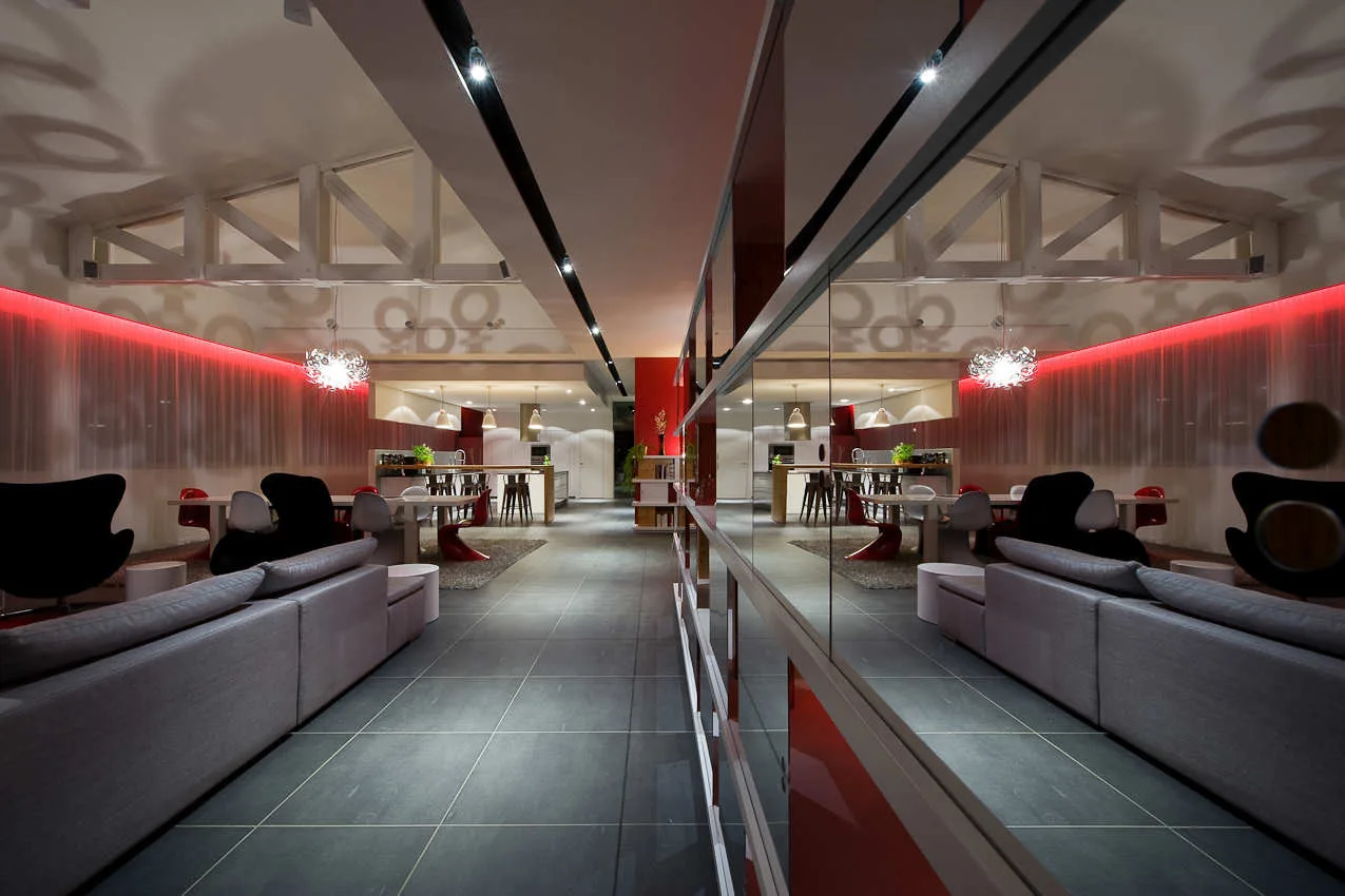 A modern open-concept room features a large grey sofa, dark tiled flooring, white ceiling beams, and a long mirrored wall reflecting the space.
