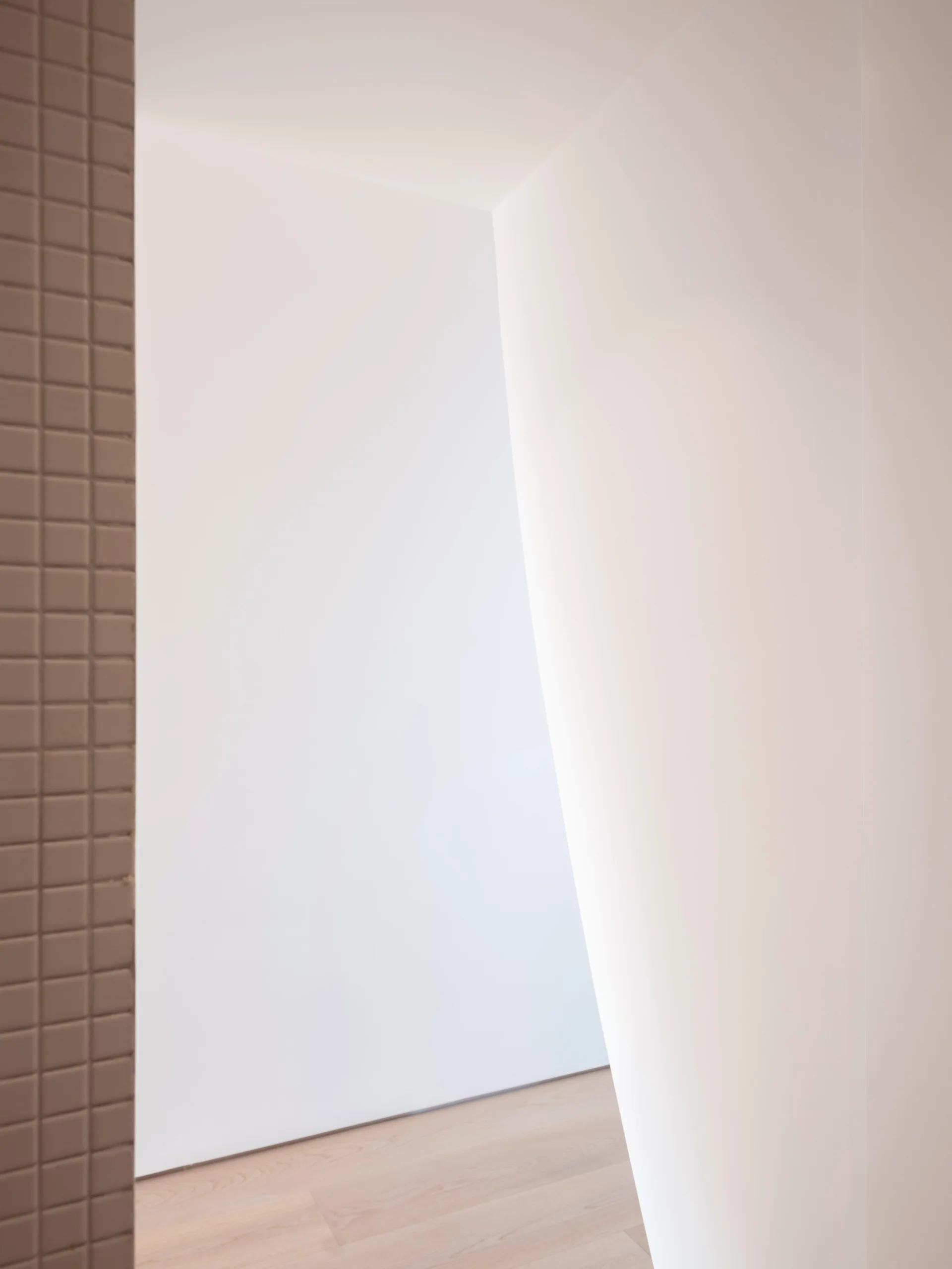 A close-up of a minimalist interior corner with plain white walls, a brown tiled column, and light wood plank flooring.