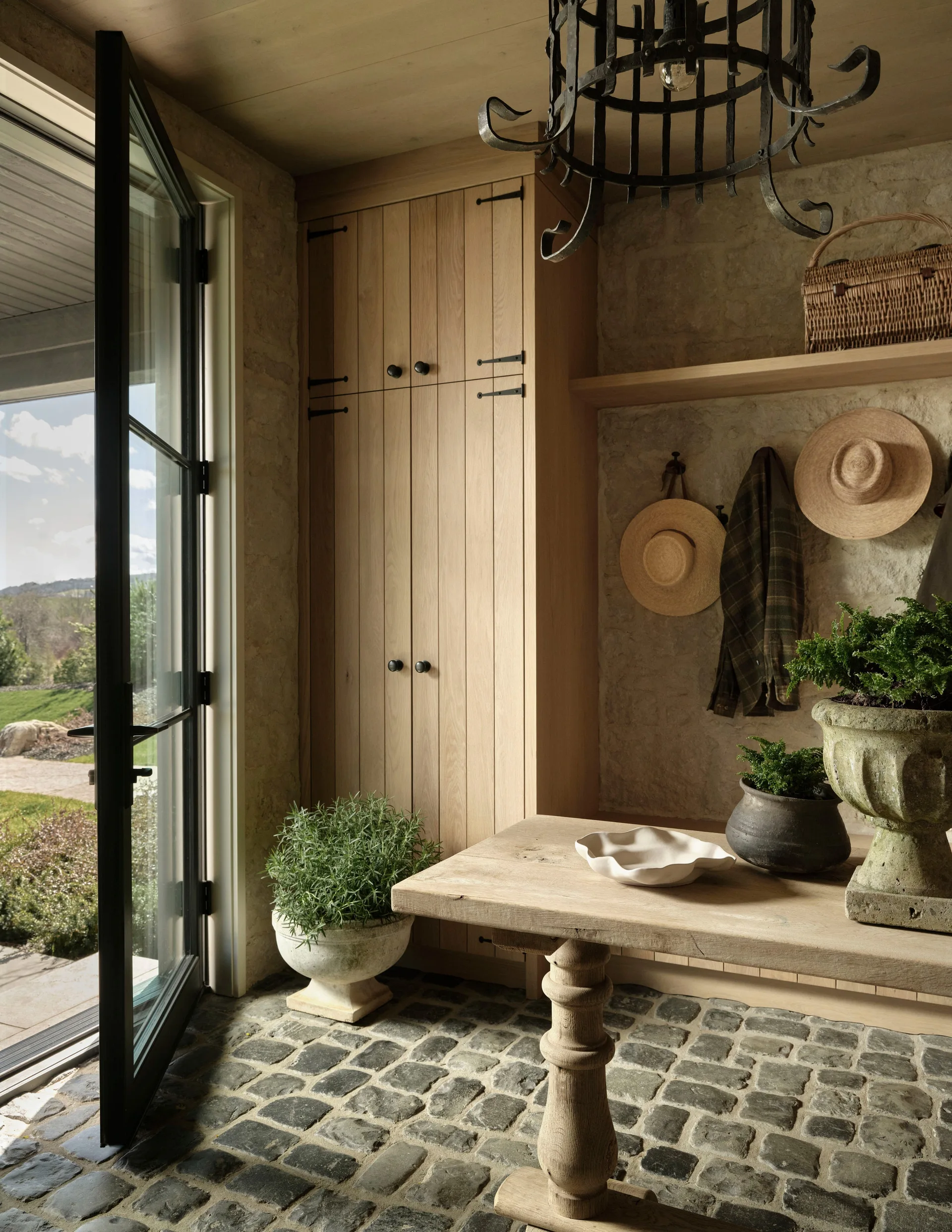 A rustic mudroom featuring irregular stone cobble flooring, tall wood cabinets, a wooden pedestal table, and an open glass door leading outside.