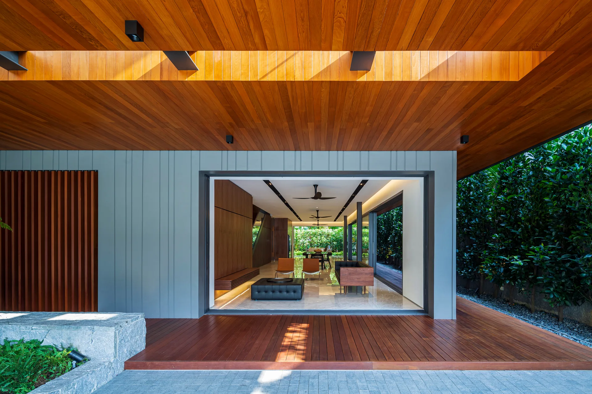 A modern living room viewed from a wooden deck, featuring a warm wood ceiling, gray vertical wall paneling, and fully open sliding glass doors.