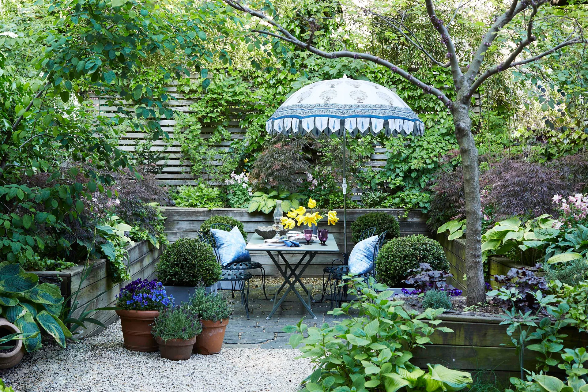 A metal dining table and chairs with blue pillows sit under a patterned umbrella on a stone patio surrounded by lush raised wooden planters.
