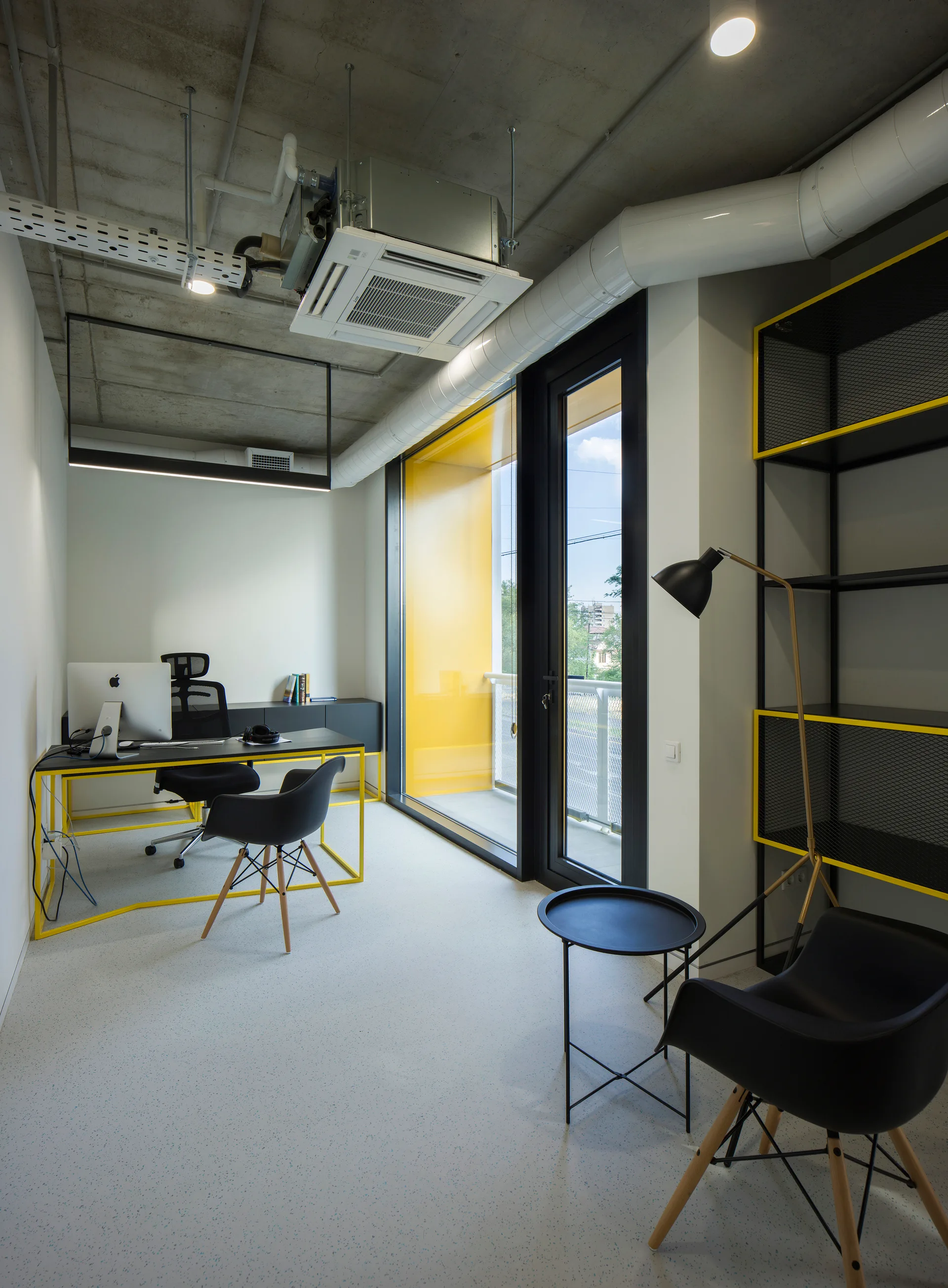 A modern industrial home office featuring a black desk with a yellow frame, black metal shelving, a large glass door, and an exposed concrete ceiling.