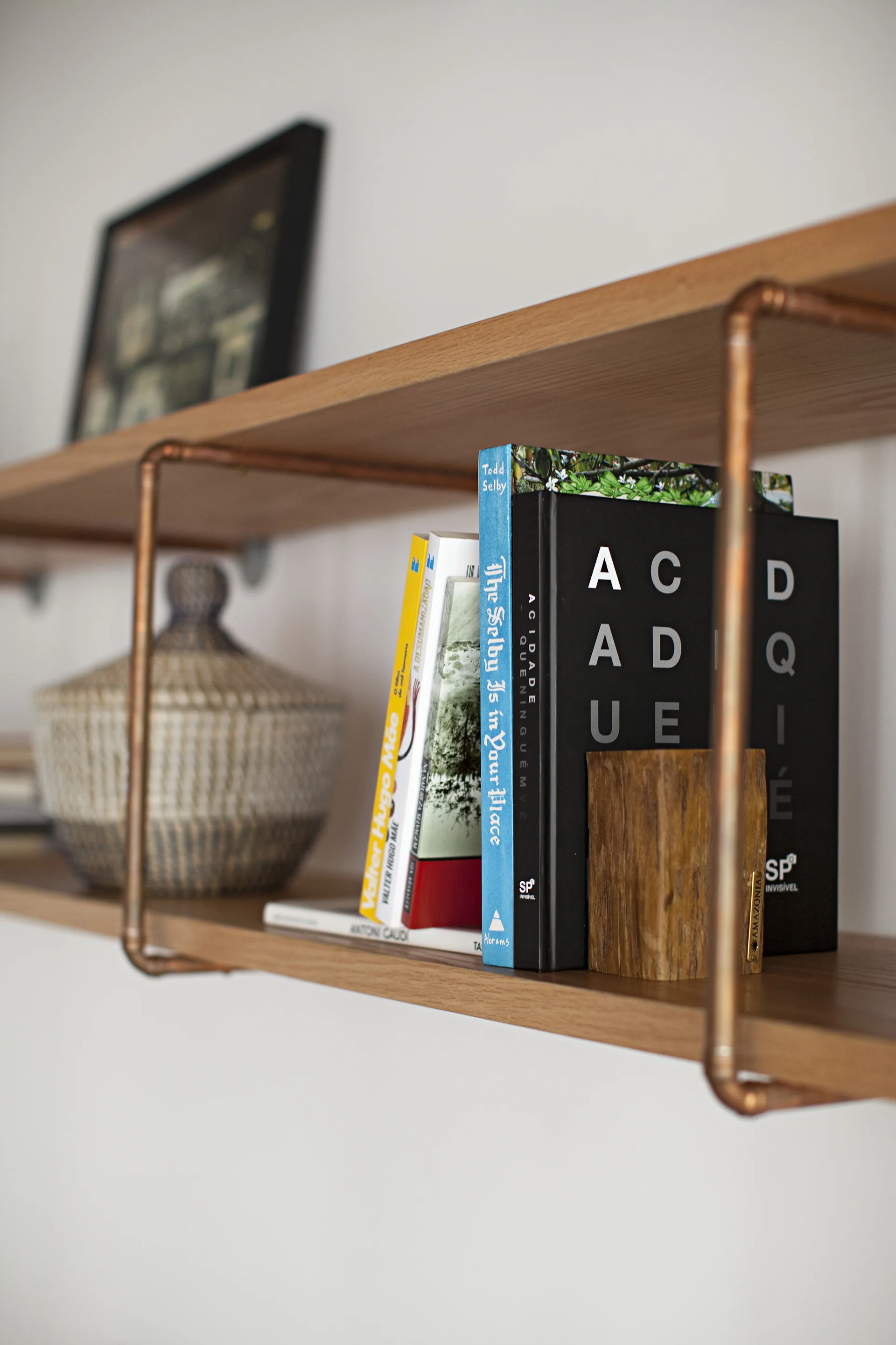 A close-up of a wooden wall shelf supported by copper pipe brackets, holding books, a lidded woven basket, and a wooden block.