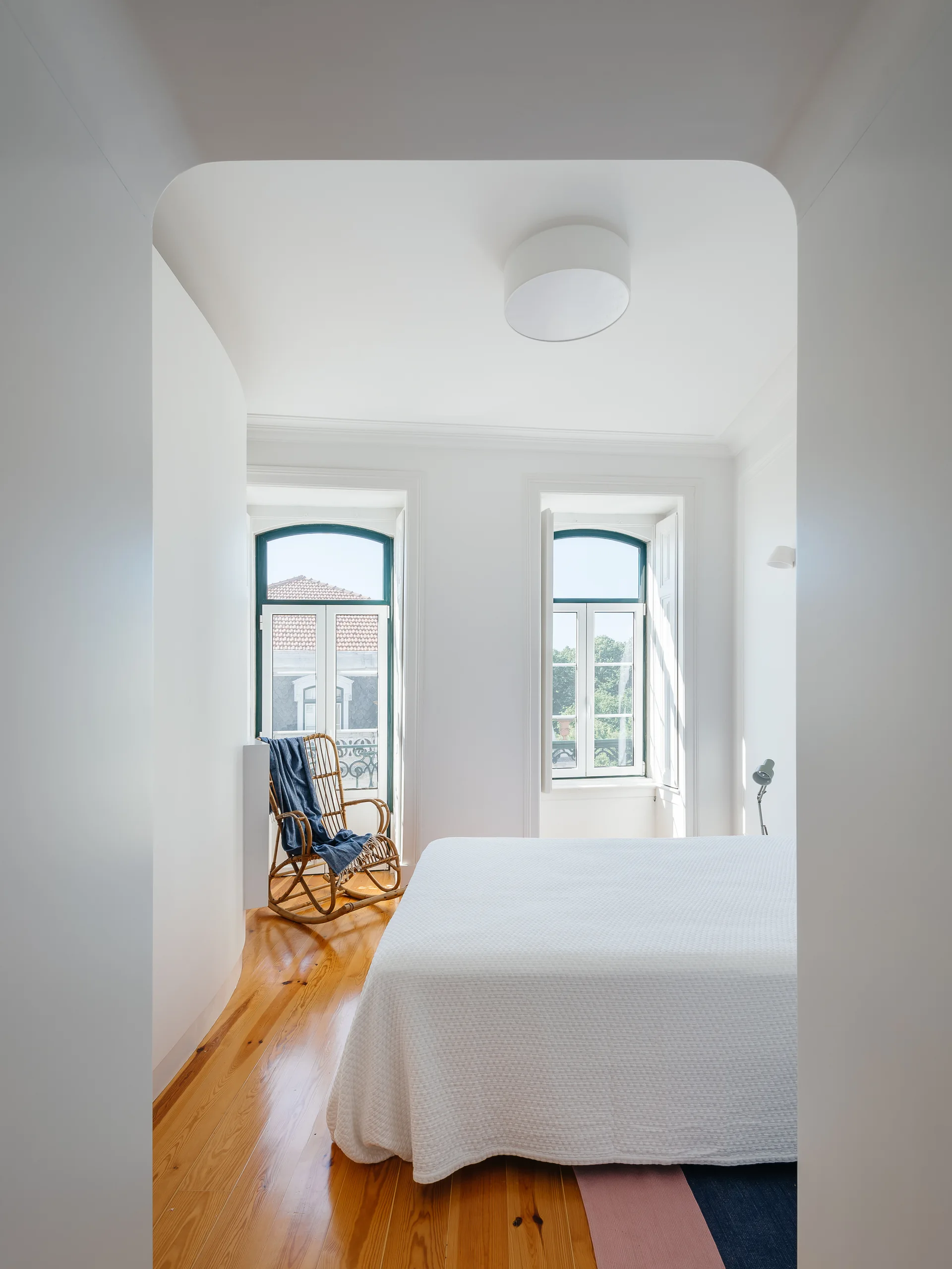 A white bedroom with glossy wood floors, viewed through a curved archway, featuring two arched windows and a rattan rocking chair.