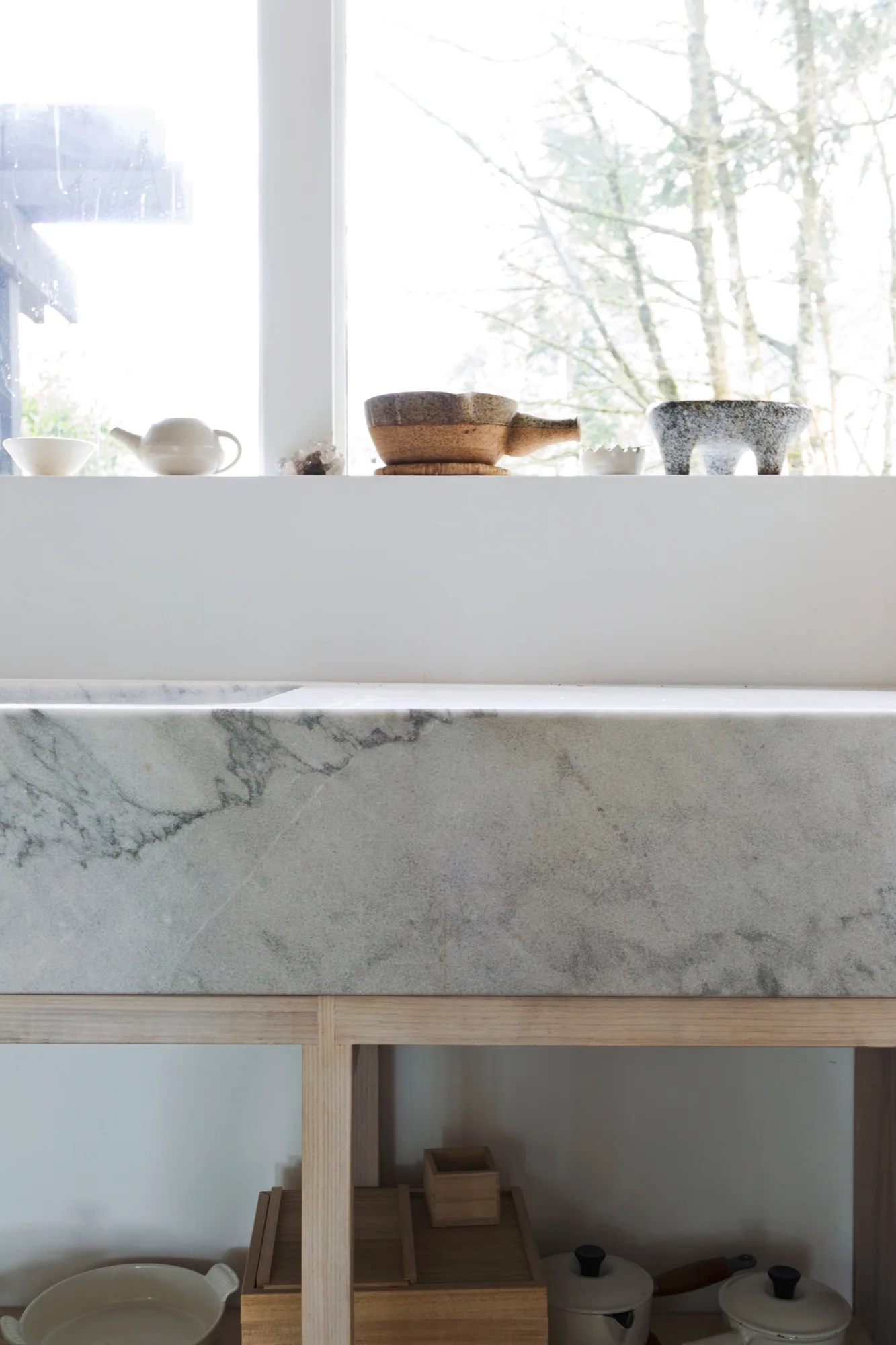 A grey veined stone basin sink rests on a light wood frame below a bright window lined with small ceramic bowls.