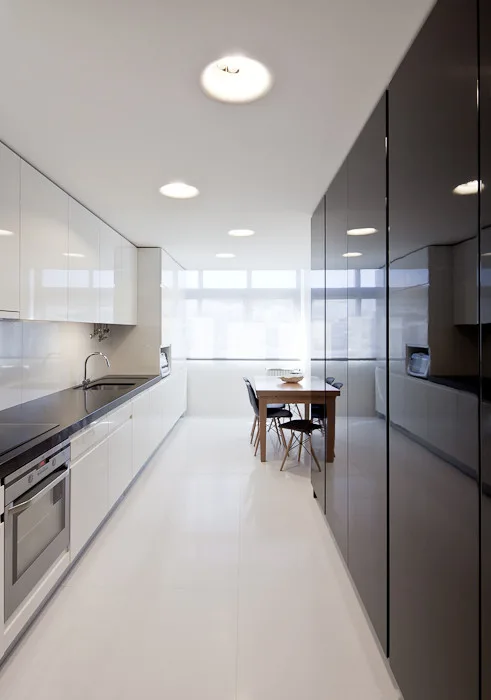 A galley kitchen featuring high-gloss white and black cabinets, a polished floor, and a small wooden dining table set by a window.