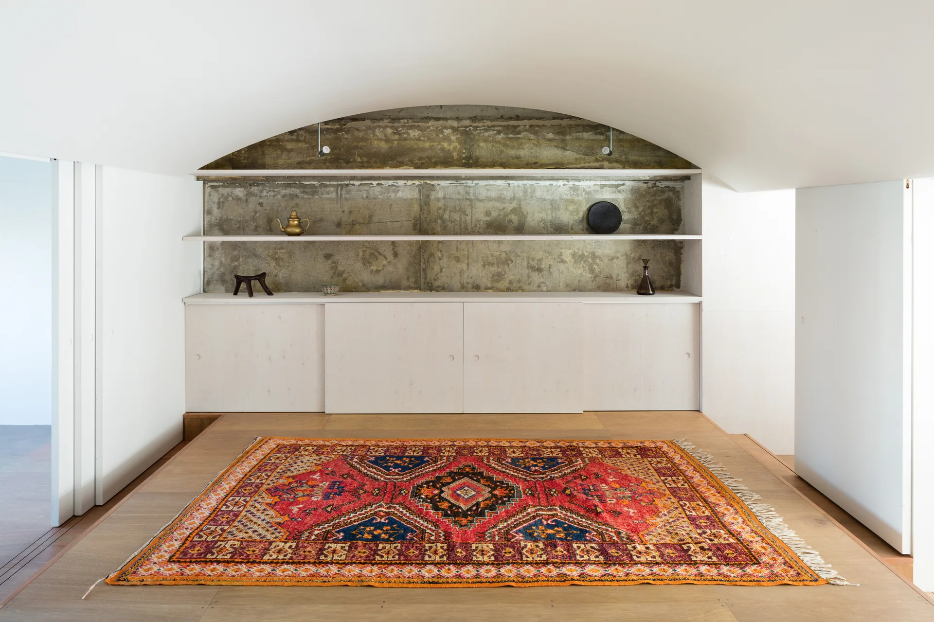 A low-ceilinged room features a vibrant red patterned rug on a light wood floor beneath an arched ceiling and wood shelves against a raw concrete wall.