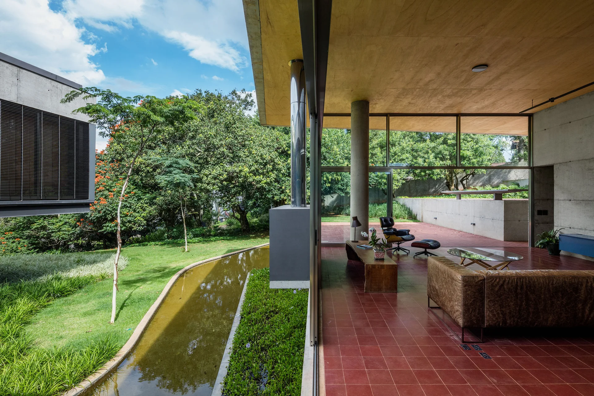A modern living room features terracotta tile floors, a brown leather sofa, and a wood ceiling with sliding glass doors opening to a lush garden.
