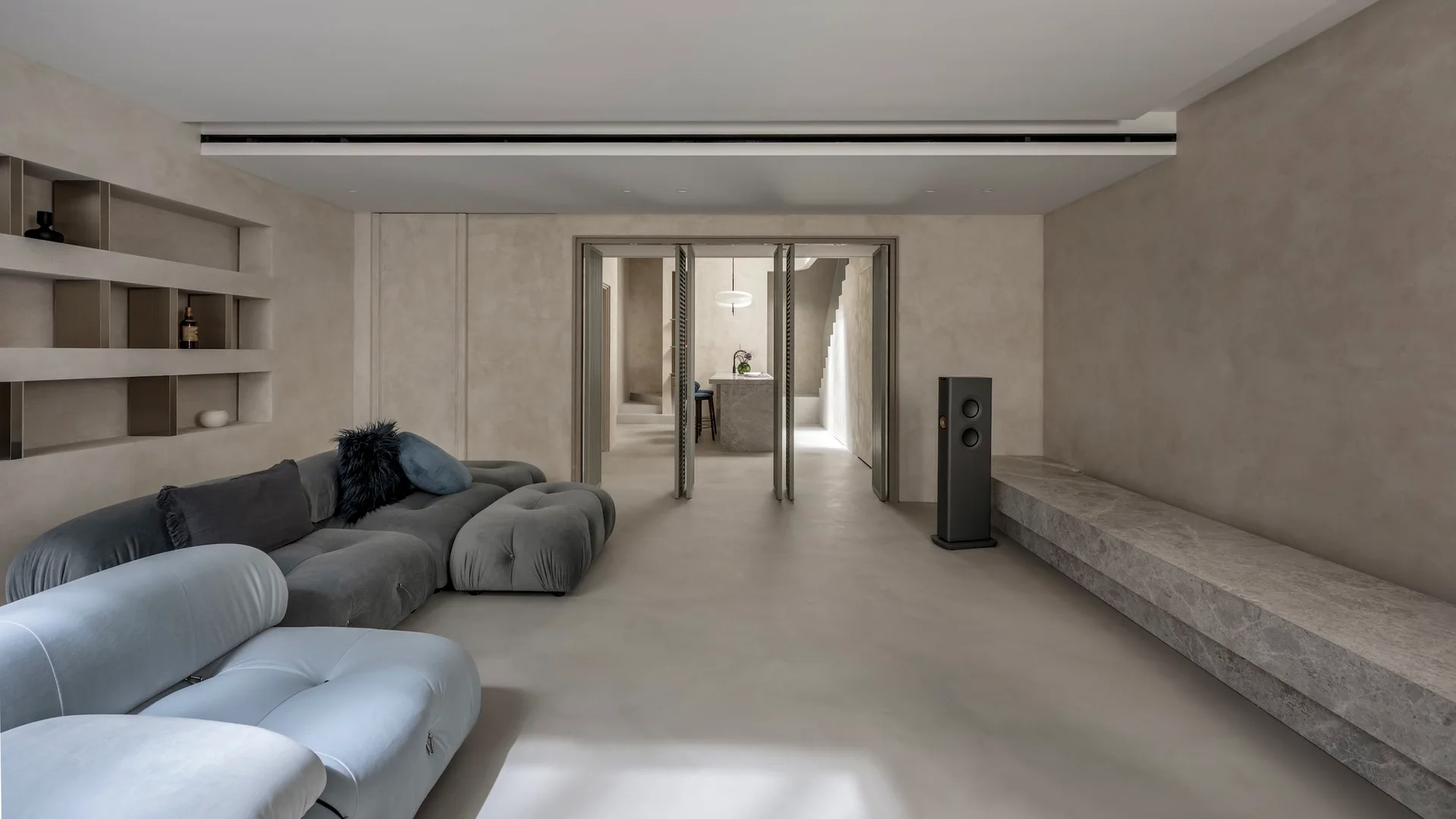 A minimalist living room features a grey modular sofa, a low stone bench, and glass bifold doors opening to an adjacent kitchen.