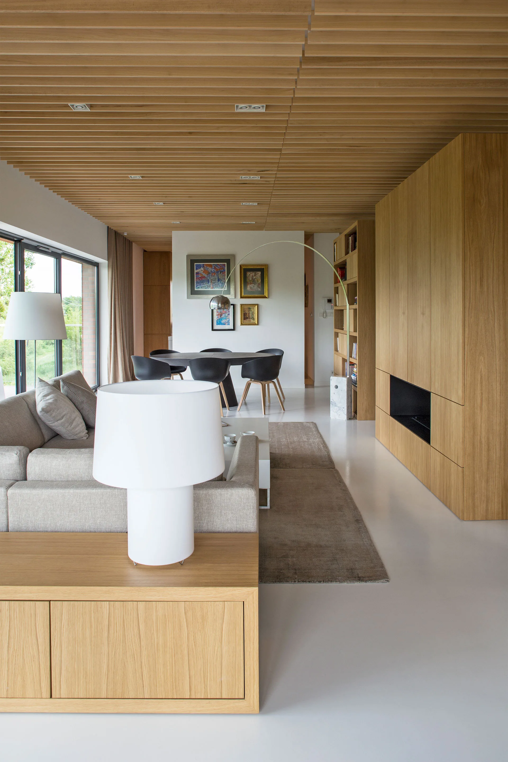 A modern open-plan living and dining space with a wood-slatted ceiling, custom light wood cabinets, and white floors.