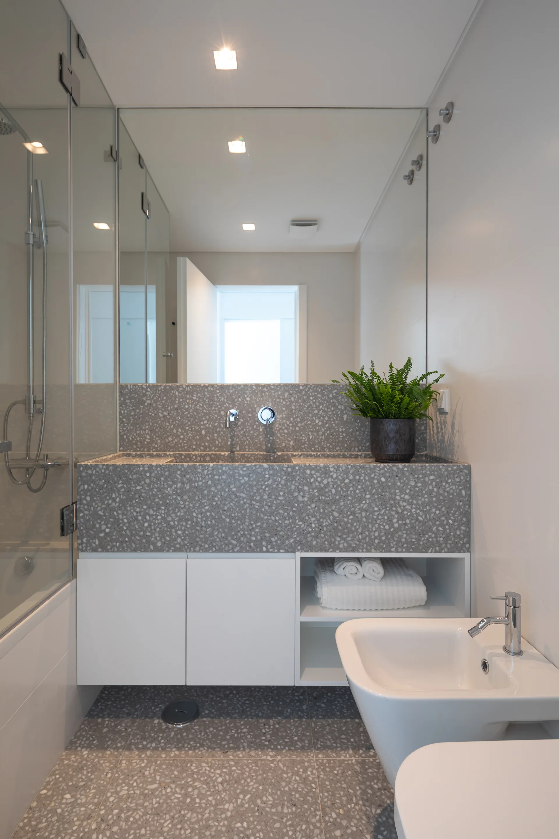 A modern bathroom features a grey terrazzo vanity and matching floor tiles, a large wall mirror, white cabinets, and a glass-enclosed shower.