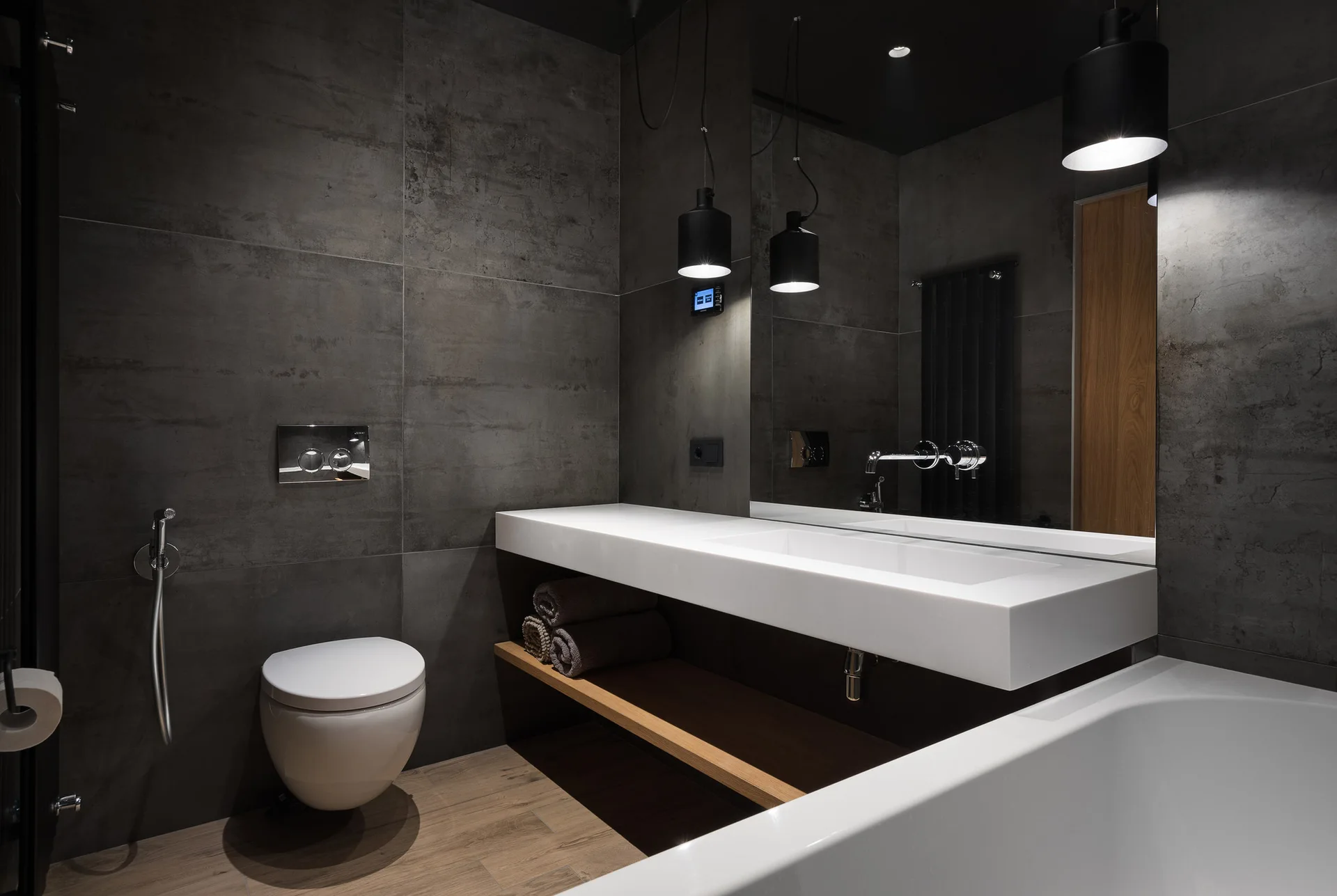 A modern bathroom featuring dark grey tiled walls, a floating white vanity with a wooden shelf, cylindrical pendant lights, and a wall-mounted toilet.