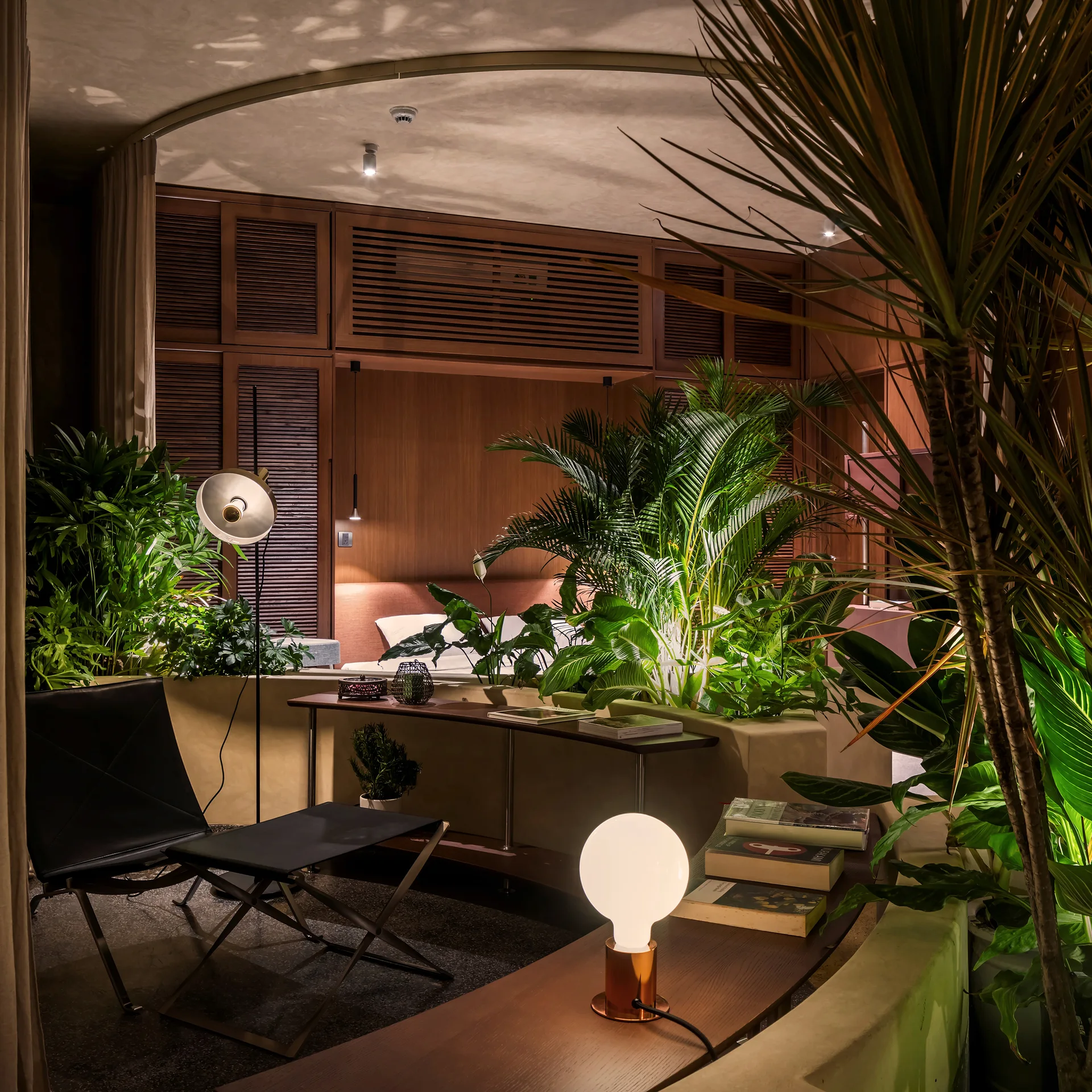 A dimly lit bedroom featuring integrated plaster planters with green plants, a black leather lounge chair, and wood-slatted wall paneling.