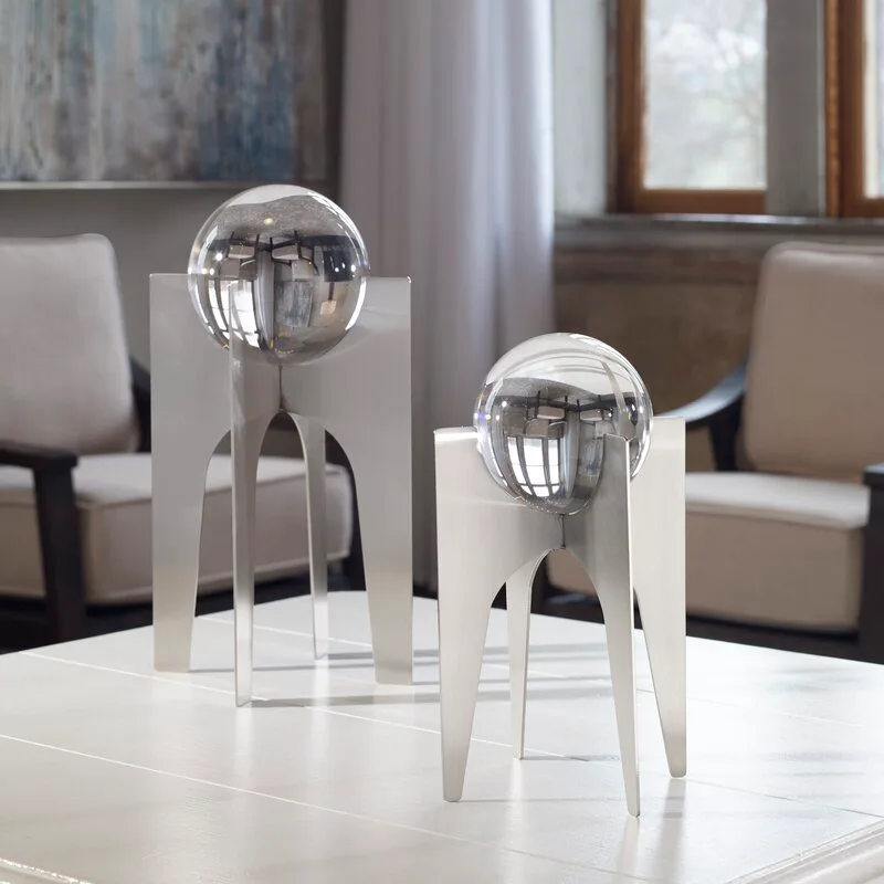 Two modern brushed metal sculptures holding reflective glass spheres sit on a white coffee table in front of blurred armchairs.