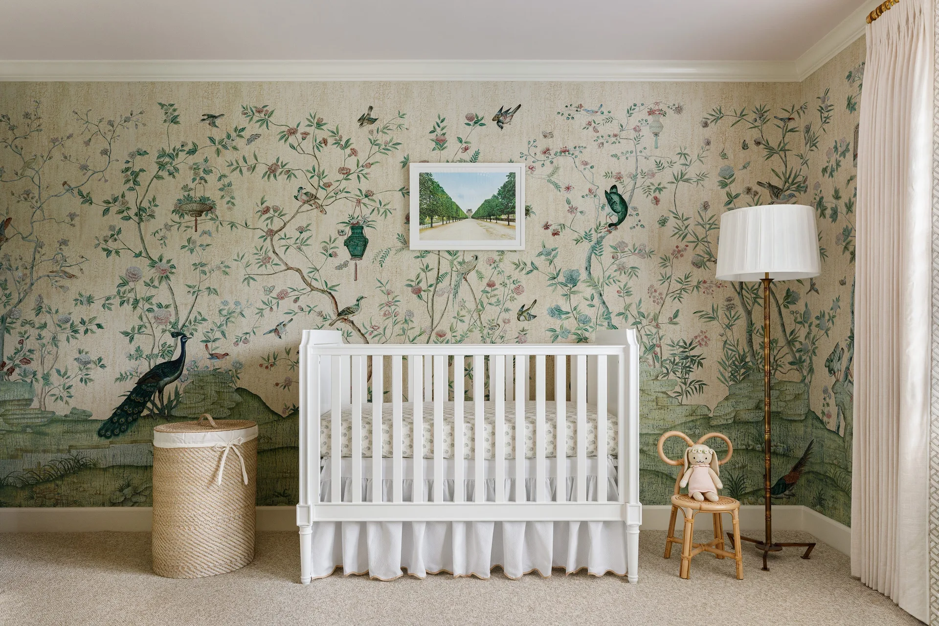 A white wooden crib sits in front of a botanical chinoiserie mural wallpaper next to a woven wicker hamper and a small rattan chair.