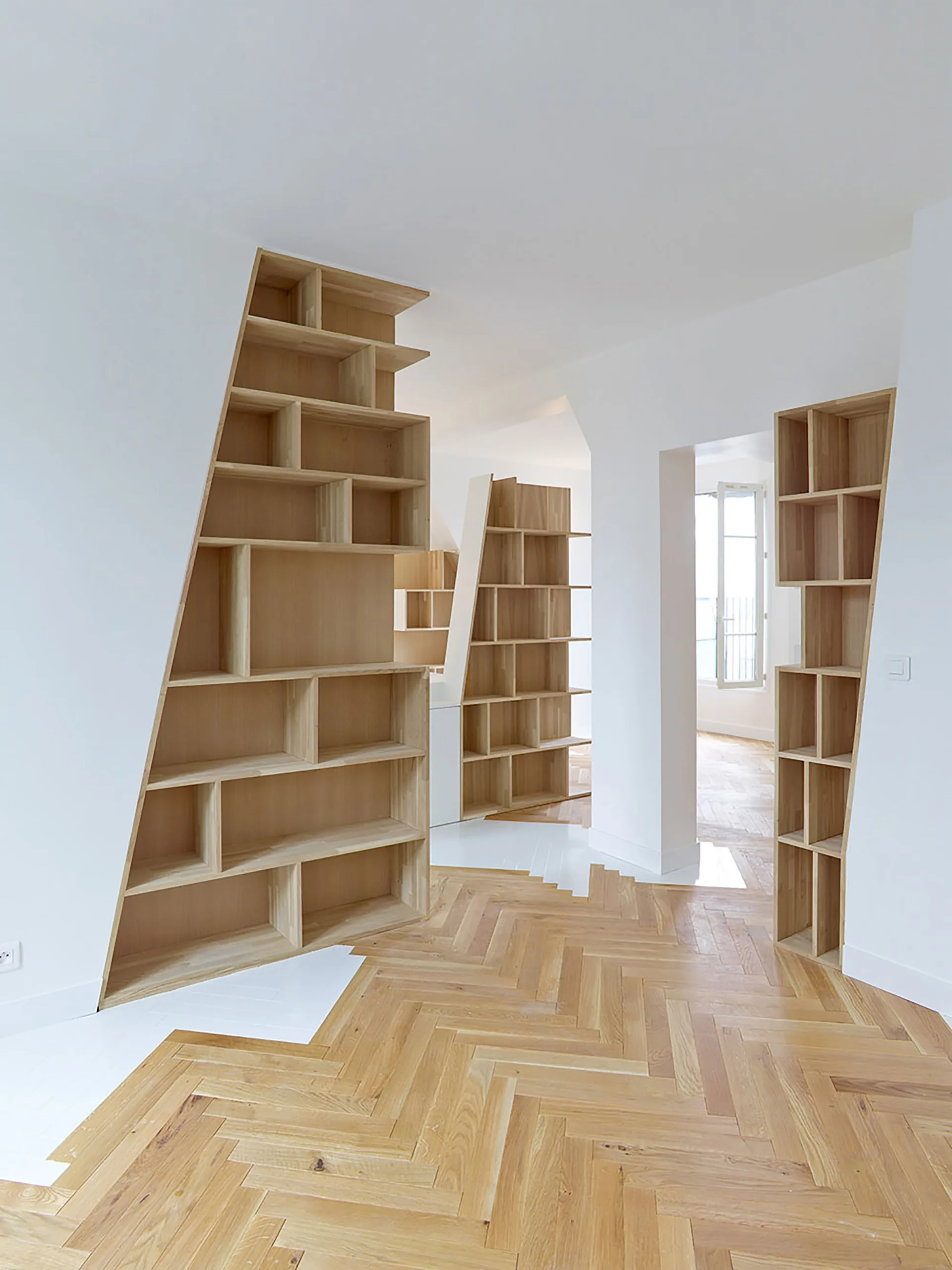 Custom-built light wood bookshelves with slanted sides stand in a minimalist room with white walls and a herringbone wood parquet floor.