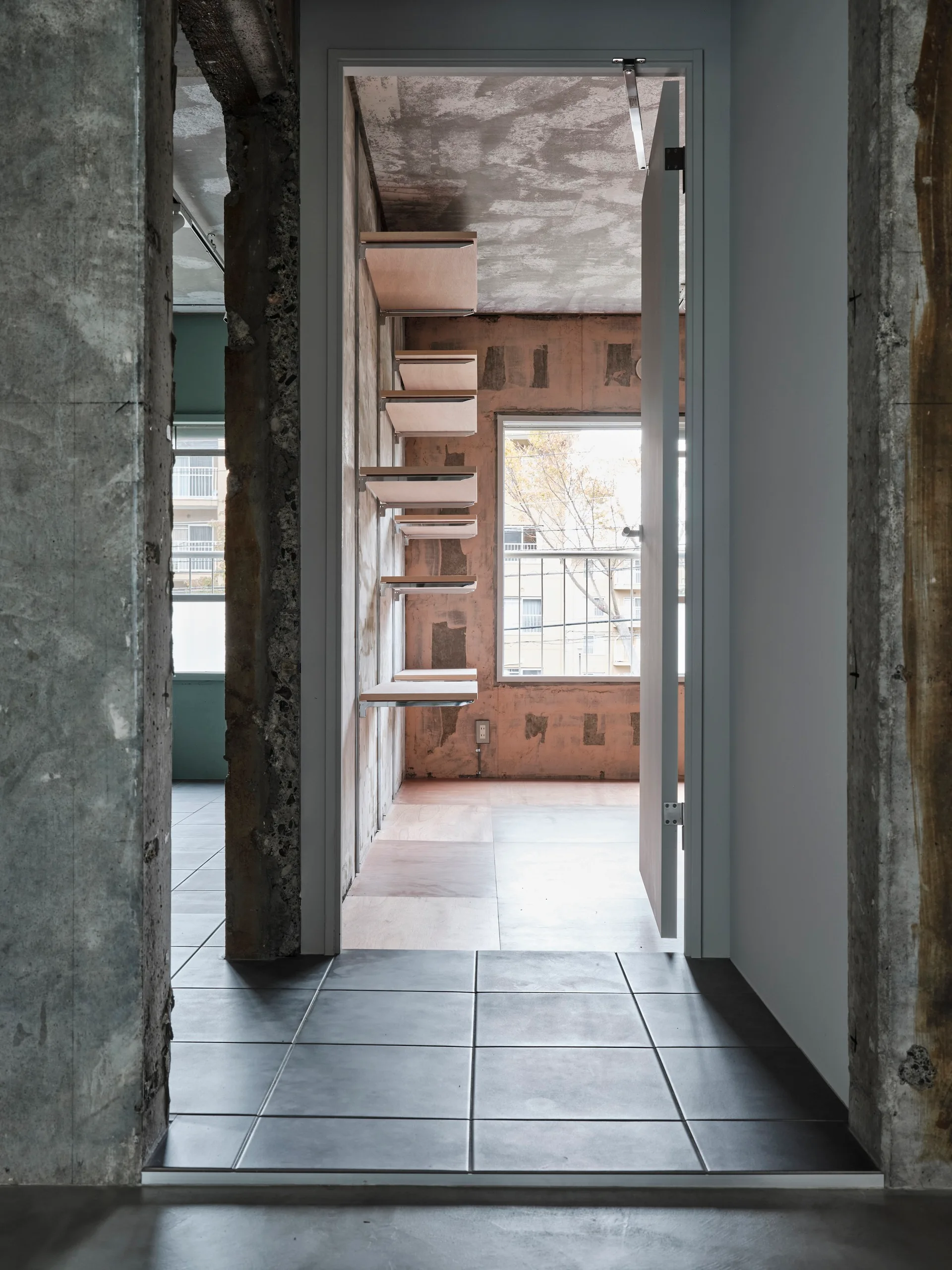A view through a doorway with grey tile flooring into an industrial room featuring exposed concrete walls and wall-mounted plywood shelves.