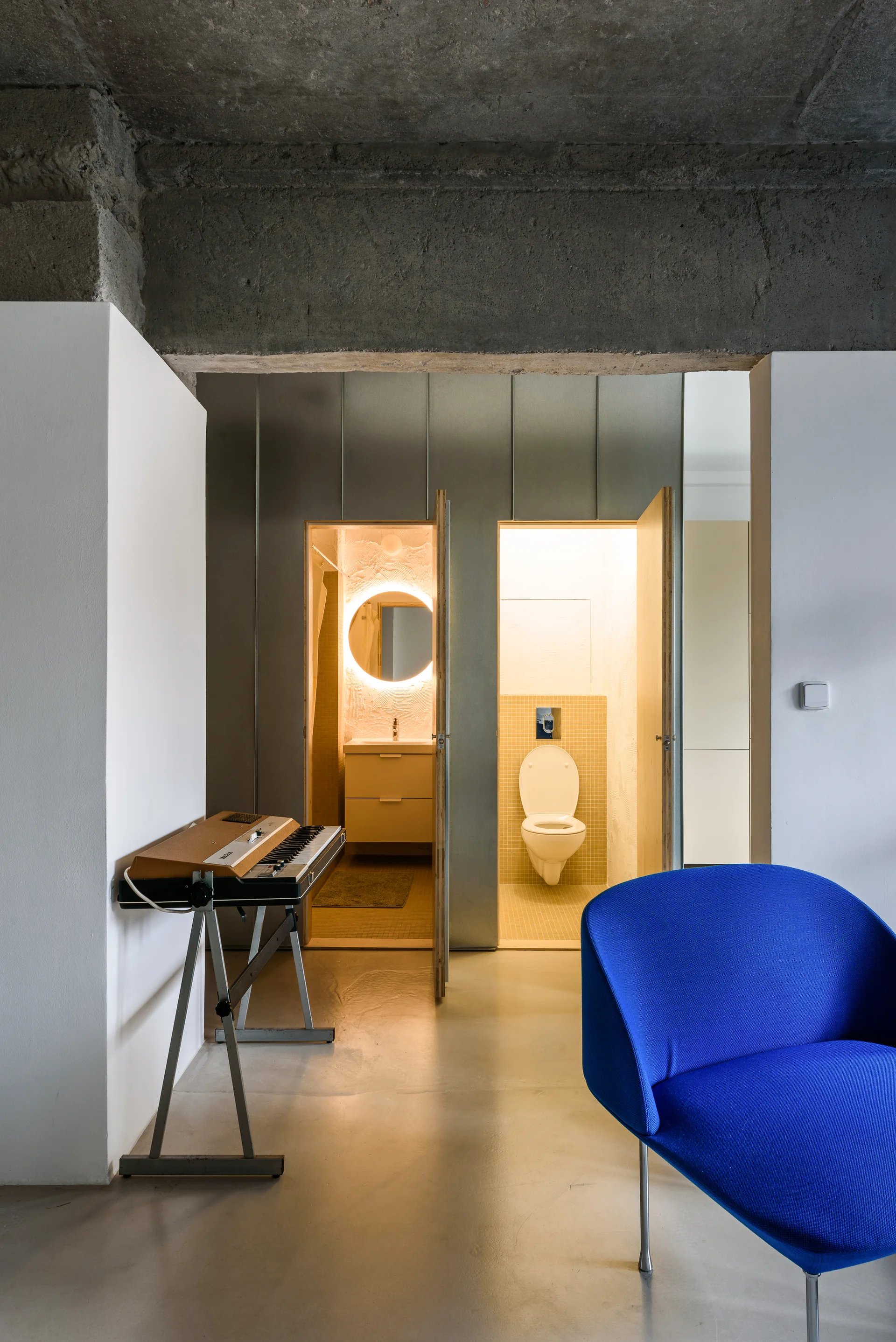 An interior space features a raw concrete ceiling, a blue armchair, and a metallic wall with dual doors opening to a sink vanity and a toilet.