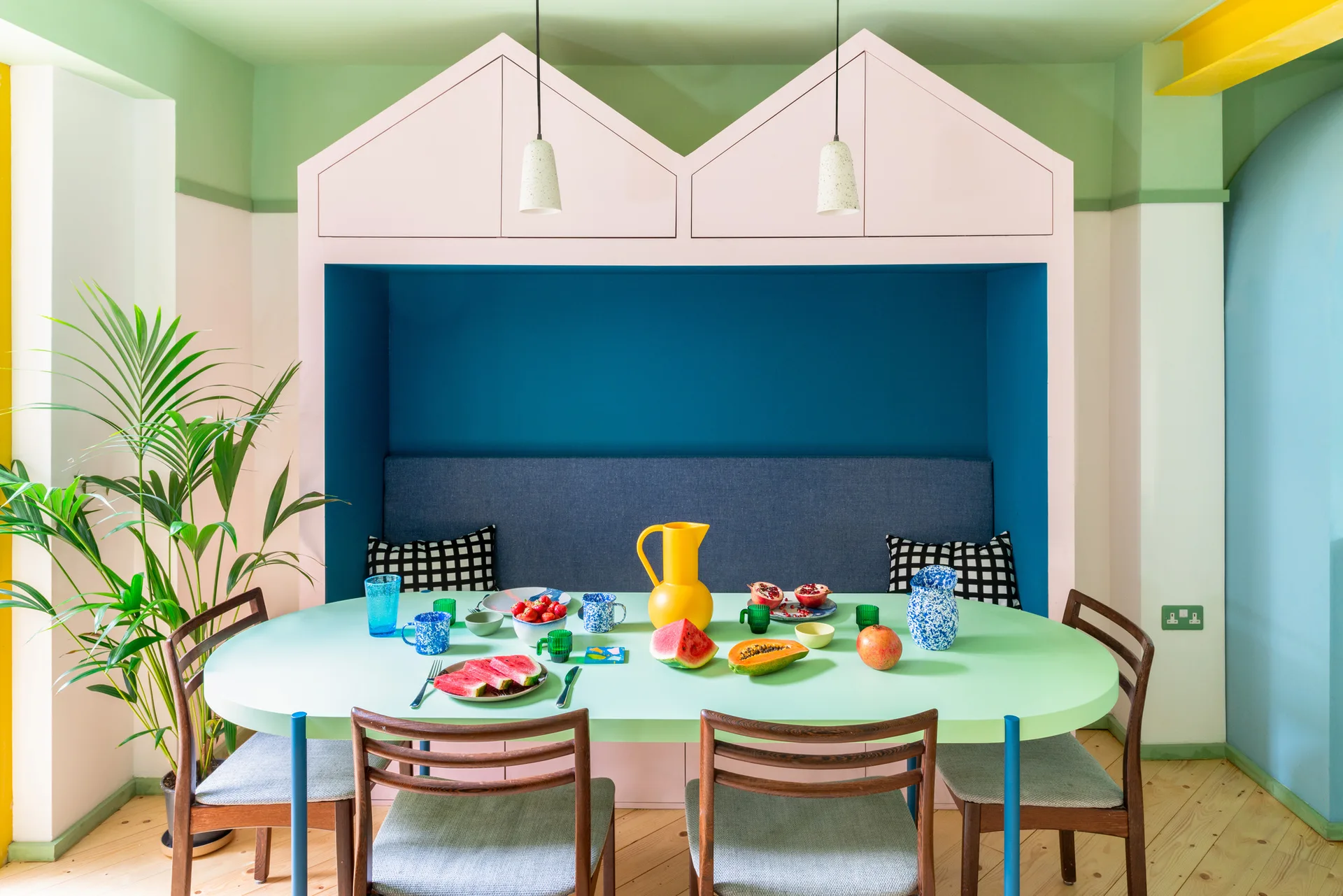 A colorful dining area features a mint green oval table, wooden chairs, and a house-shaped built-in pink and blue bench alcove.
