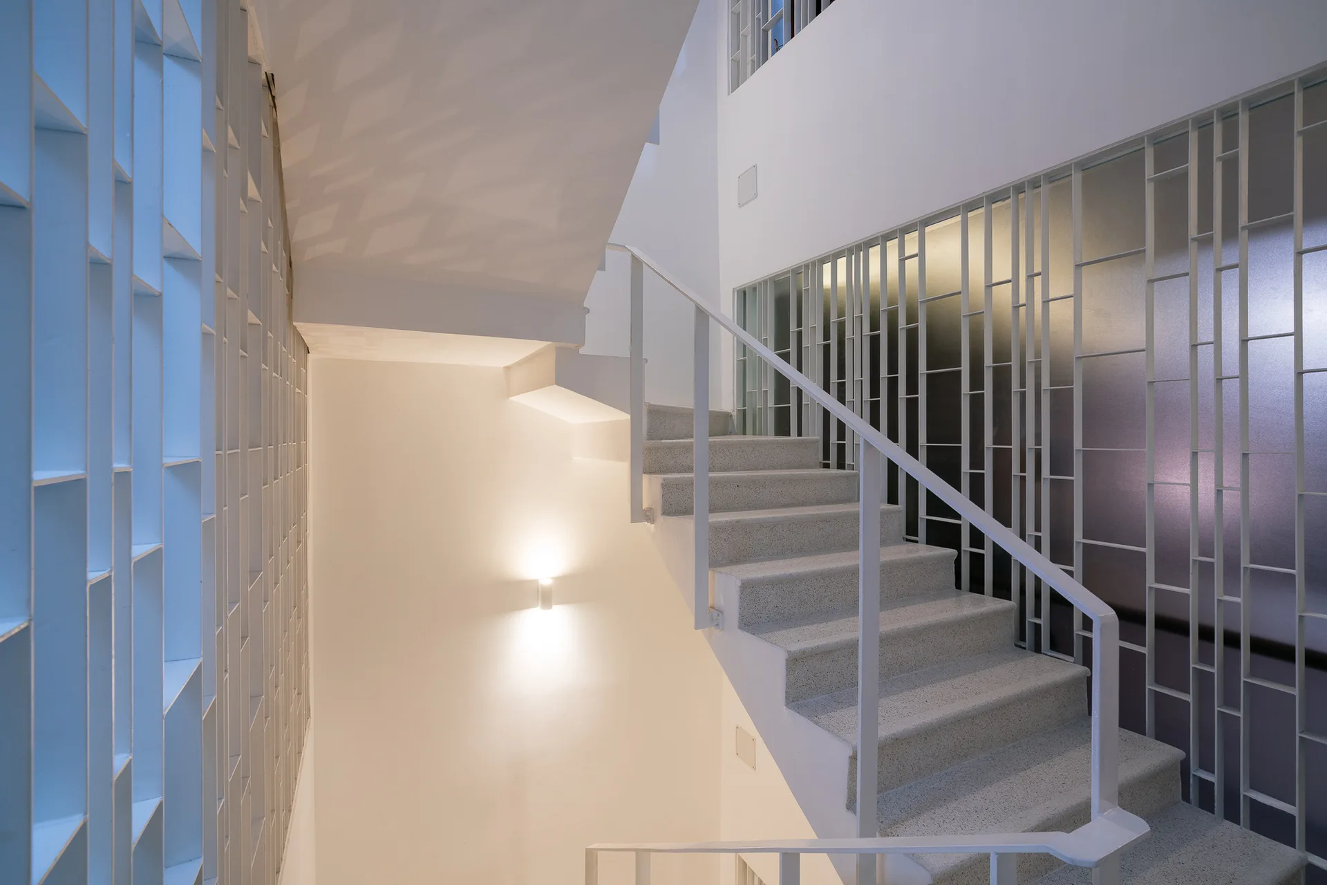 A modernist white staircase with stone-textured steps, a metal railing, and geometric patterned screen partitions illuminated by a soft wall sconce.