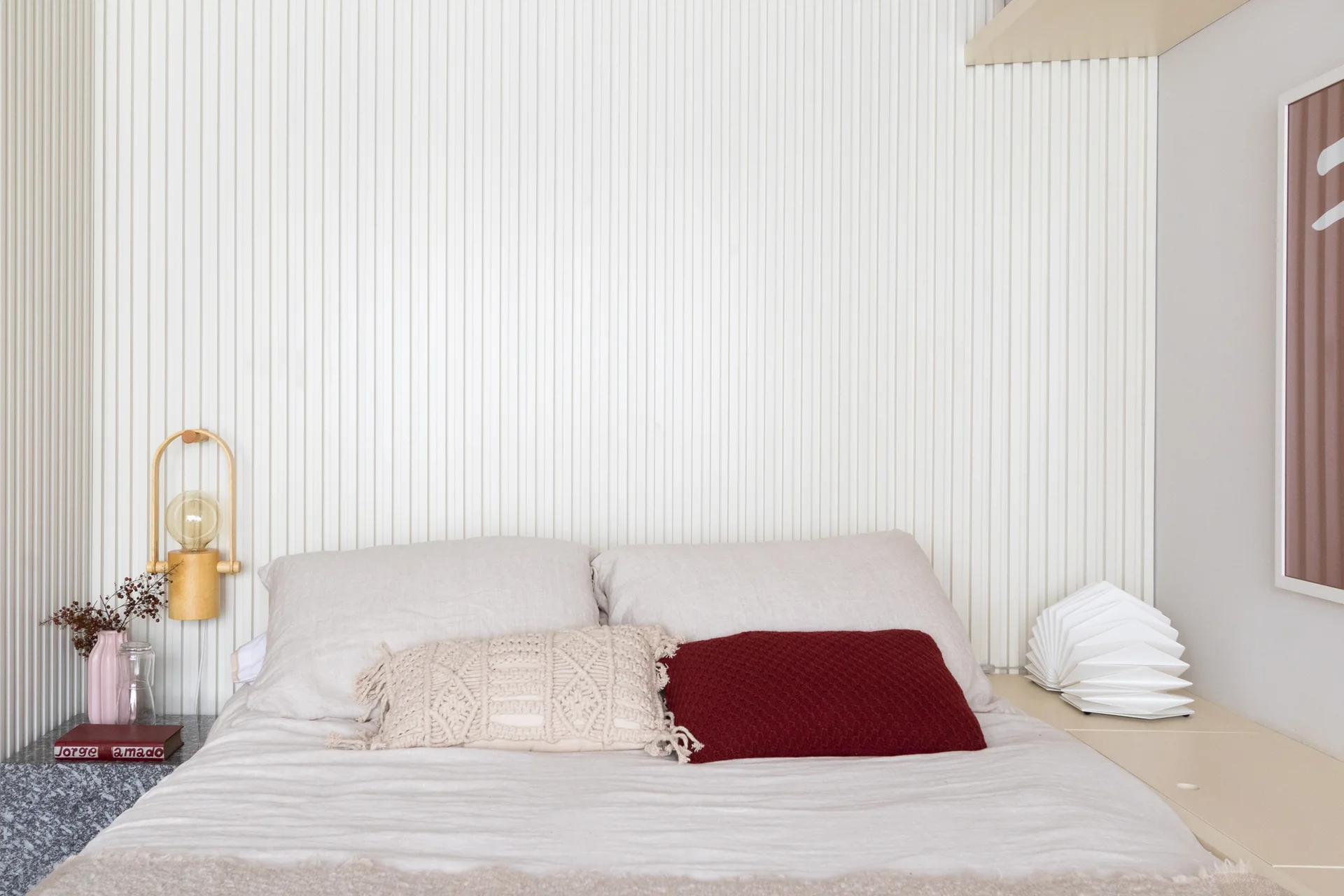 A bed with neutral linen bedding and a red throw pillow sits against a wall covered in white vertical wood slats.