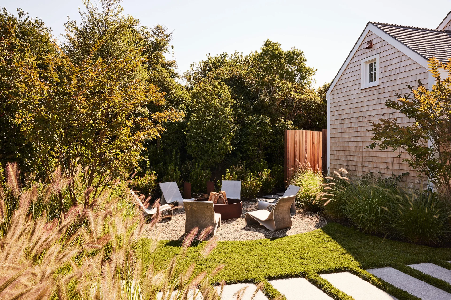 Modern curved lounge chairs surround a rusty metal fire pit on a circular gravel patio next to a shingle-sided house.