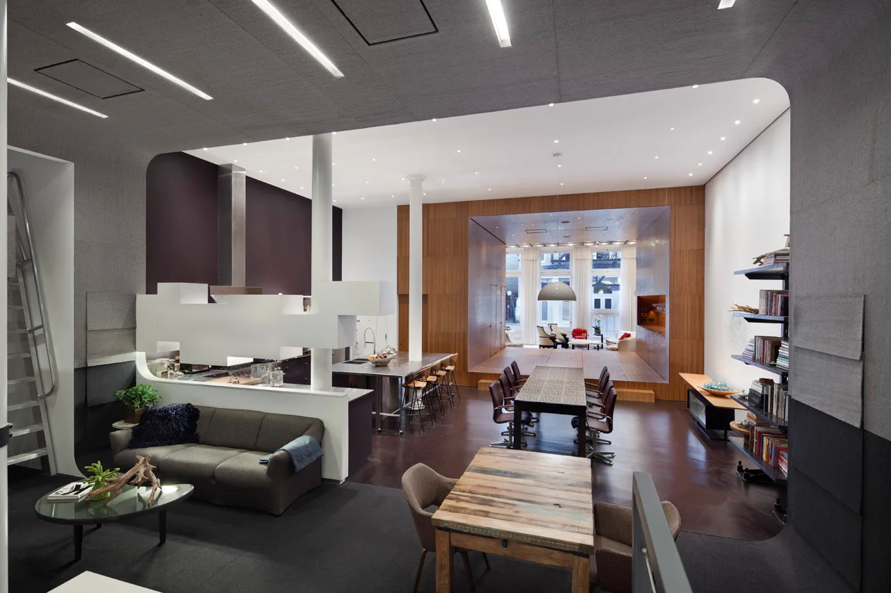 An open-plan loft features a lower seating area, a long dining table, and a raised wood-paneled platform framing a back lounge.
