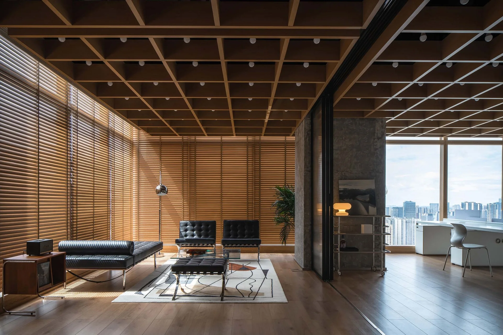 Black tufted leather chairs and a daybed sit under a wooden coffered ceiling next to large windows with wood horizontal blinds.