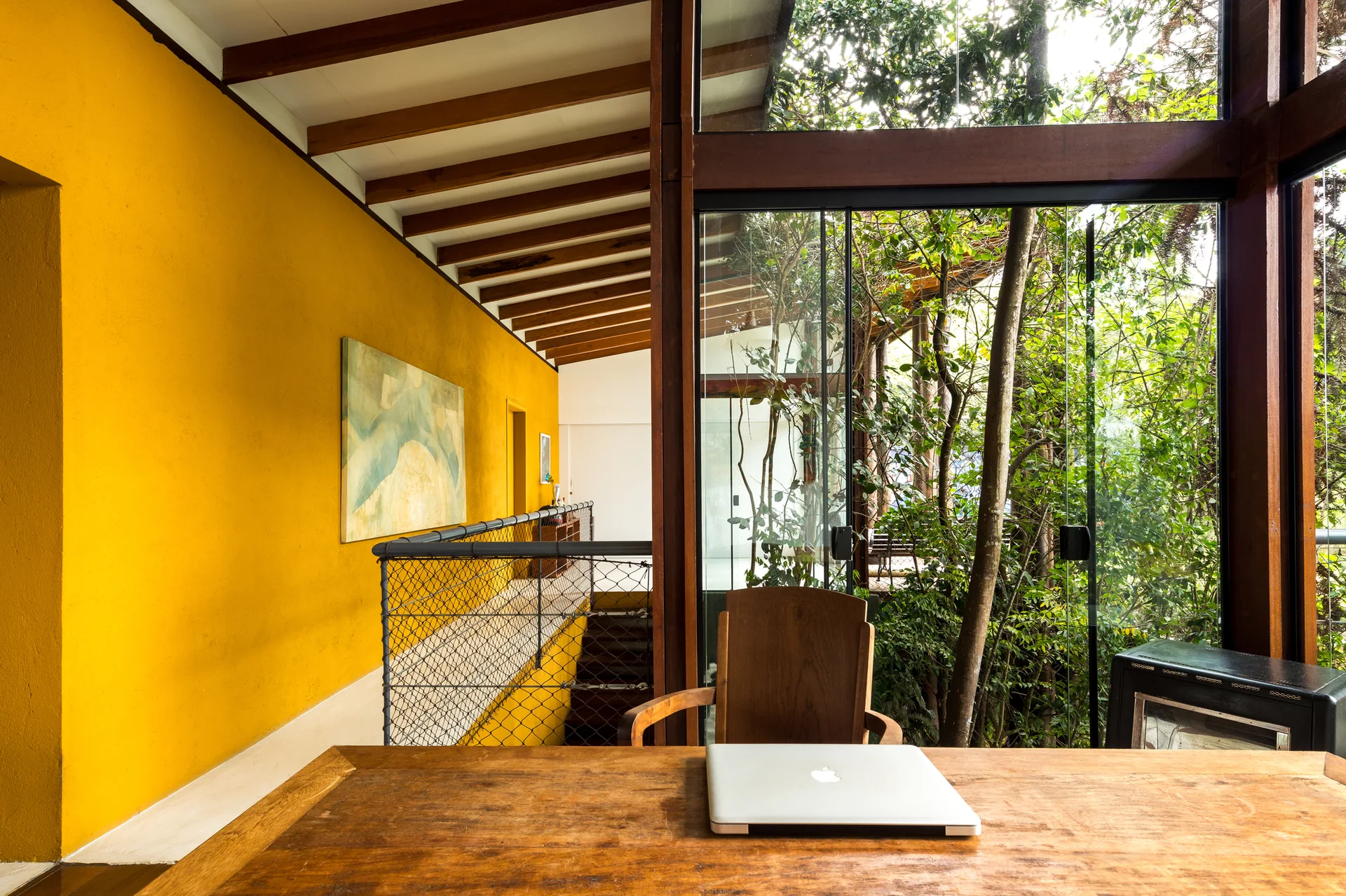 A home office features a wooden desk with a laptop, exposed ceiling beams, and a bold yellow wall next to large windows overlooking trees.