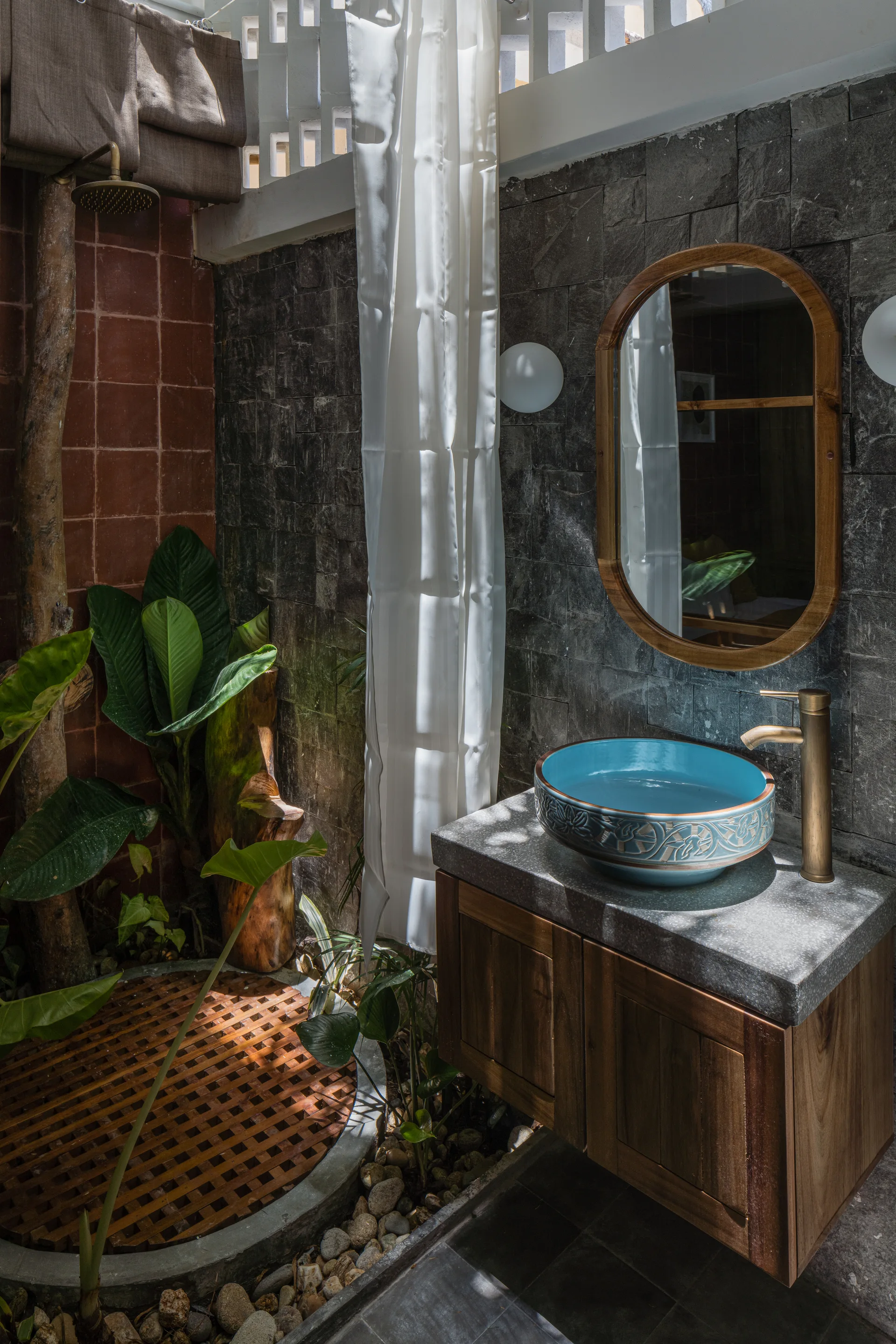 An indoor-outdoor tropical bathroom features a dark slate tile wall, a blue patterned vessel sink on a wooden vanity, and a wooden slat shower floor.