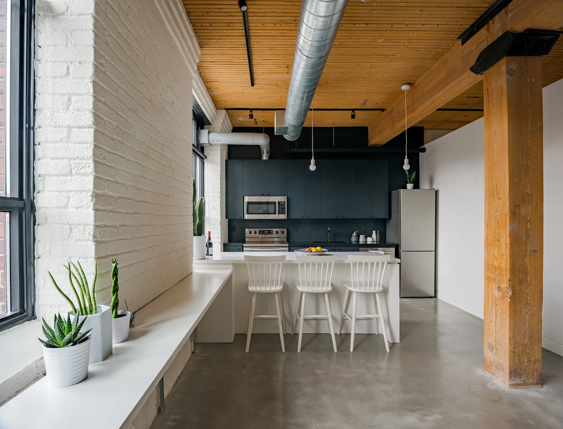 An industrial kitchen with matte black cabinetry, a white island with bar stools, painted brick walls, and exposed wooden ceiling beams.