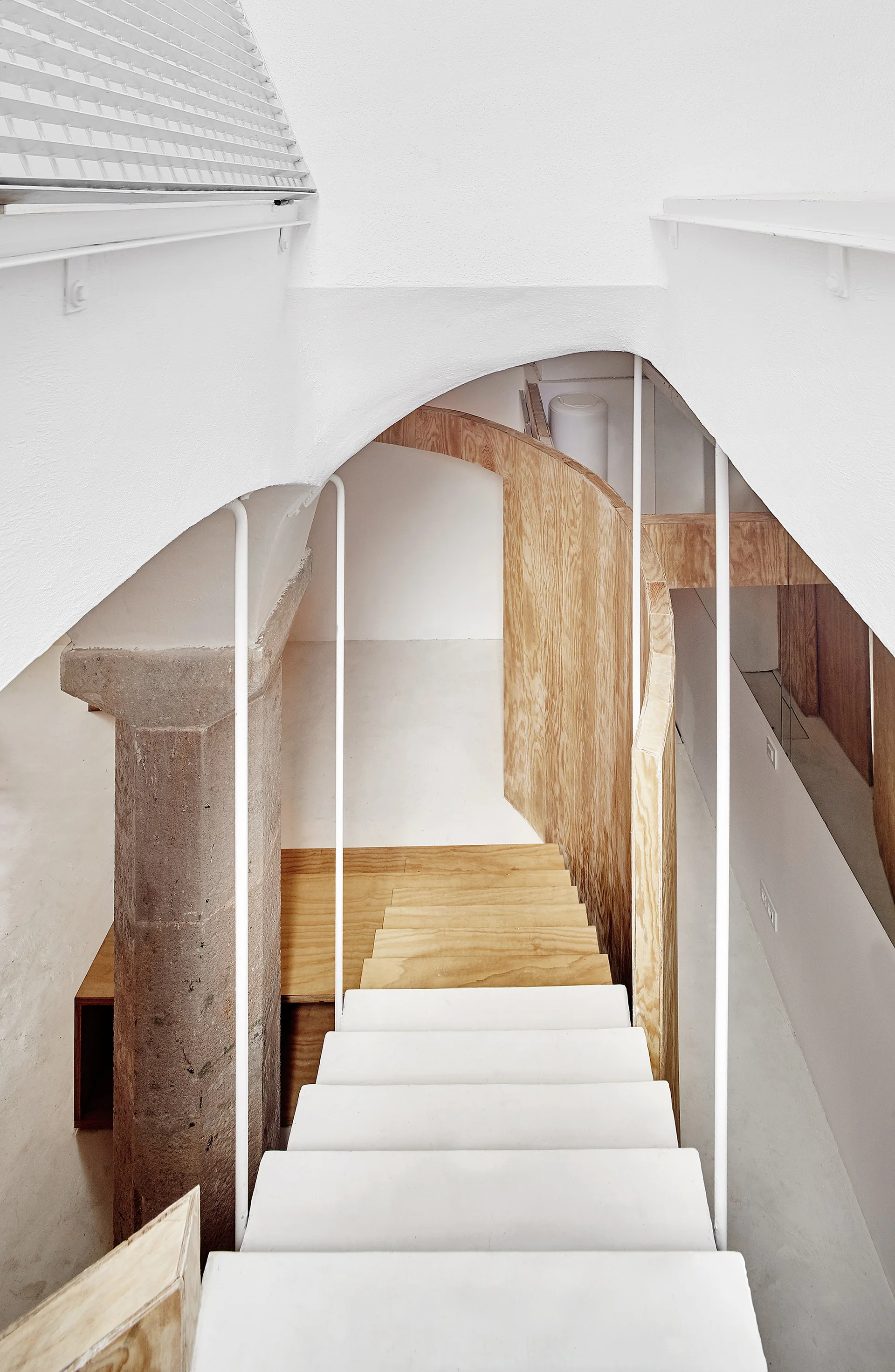A high-angle view of white modern stairs descending past a rustic stone column and a curved natural wood partition.