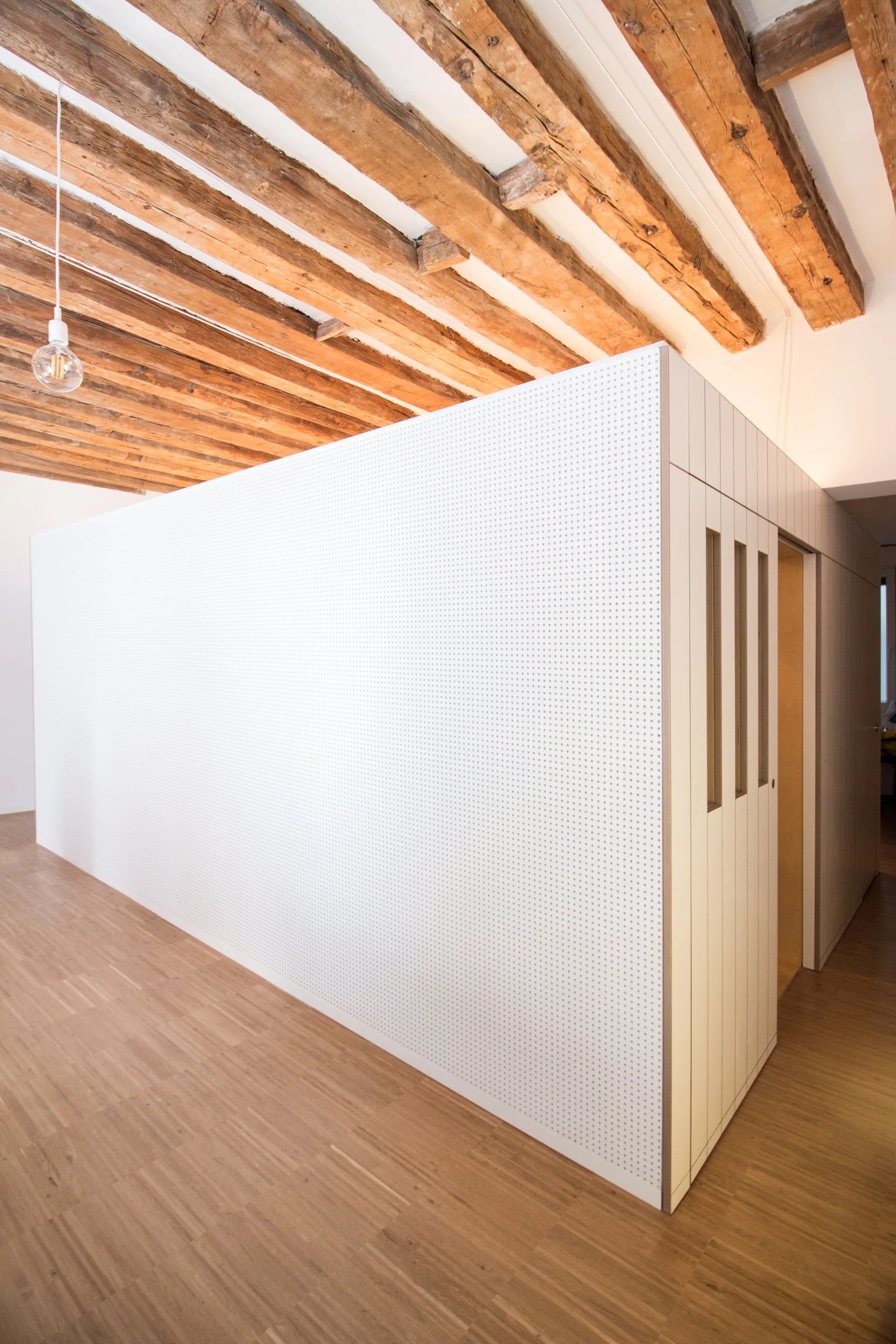 A large white pegboard room divider with an integrated slatted door stands beneath a rustic ceiling with exposed wooden beams.