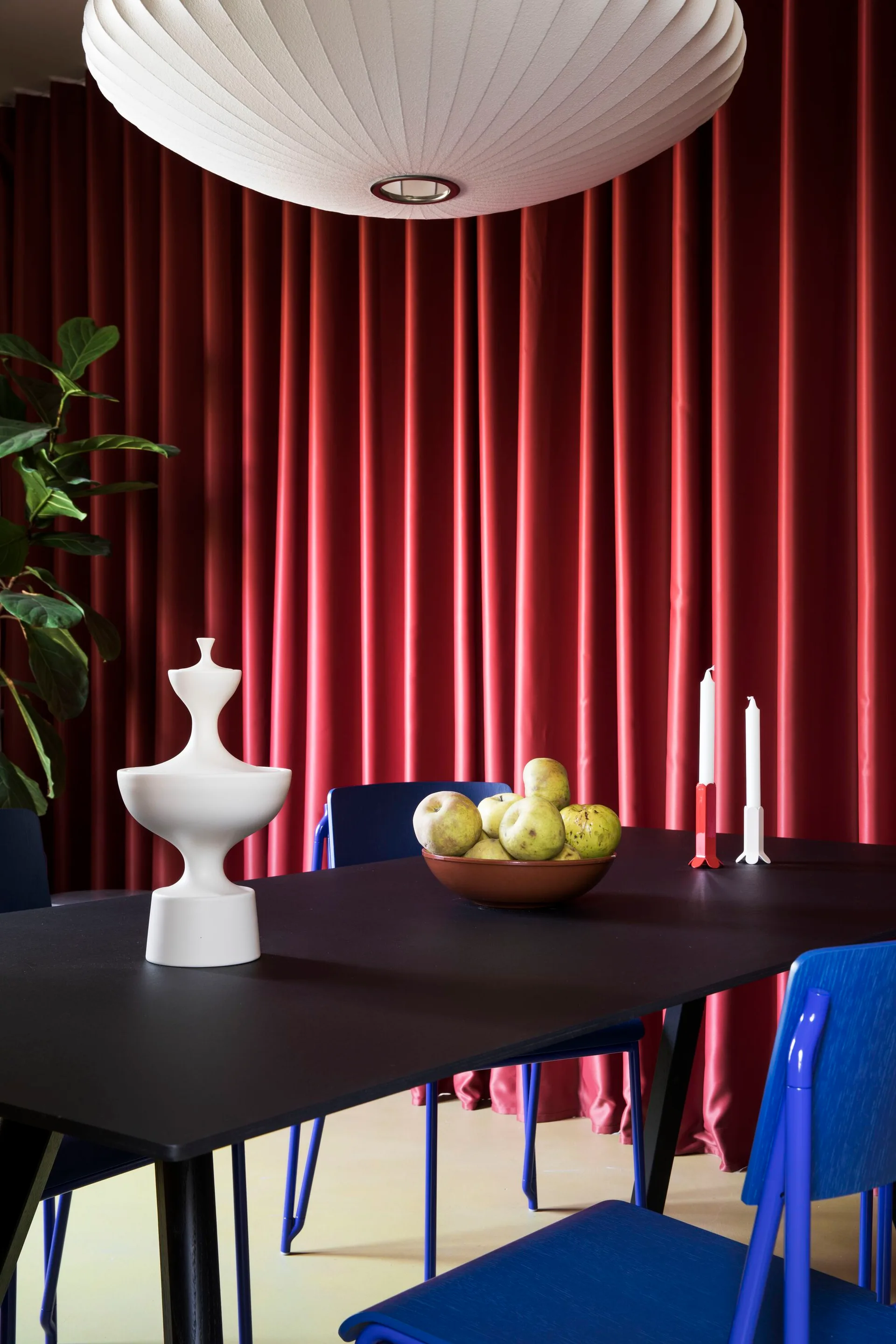A black dining table with blue wooden chairs stands before deep red pleated curtains under a large white pleated pendant light.