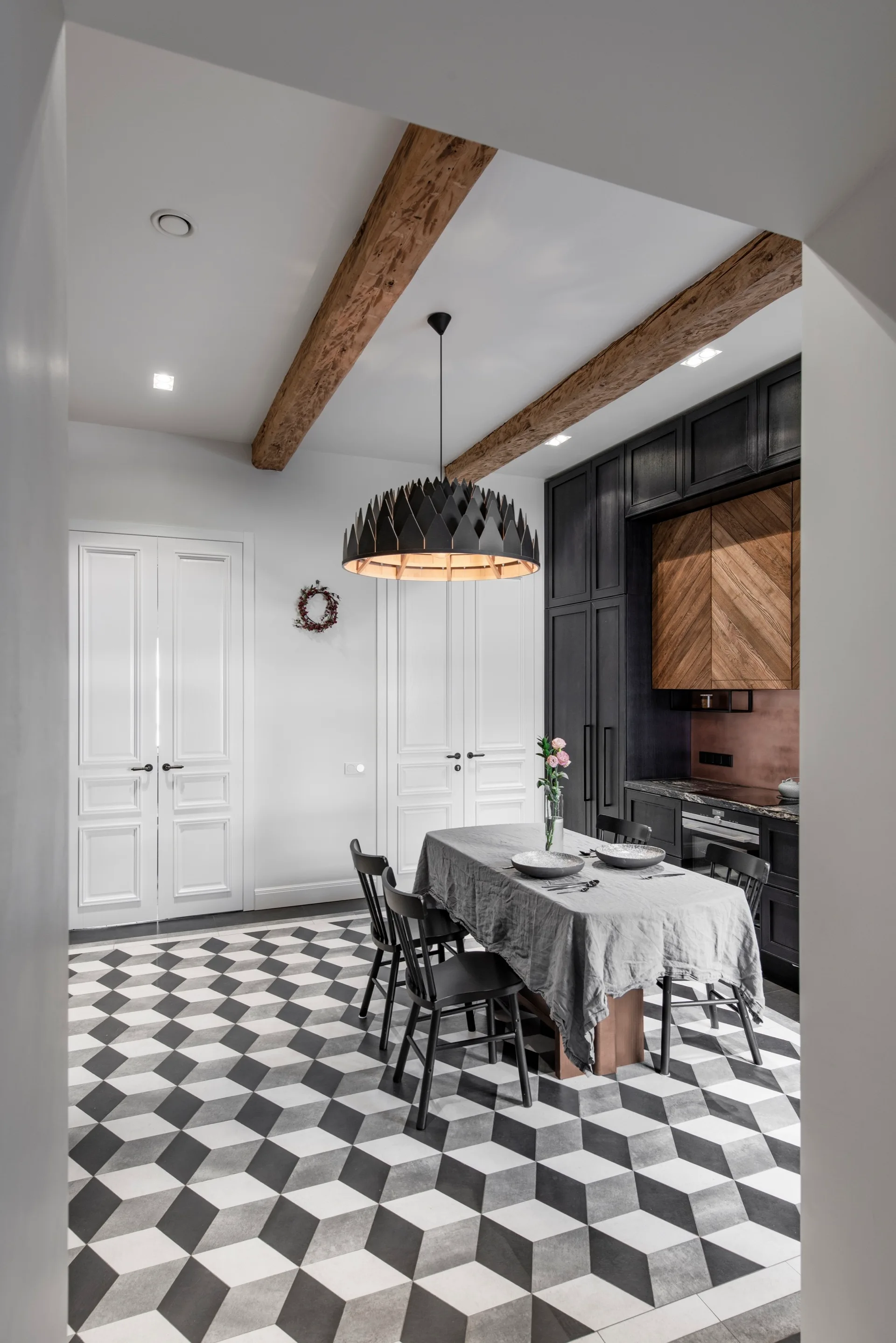 A kitchen and dining space features a black and white geometric tiled floor, a dining table with grey cloth, dark cabinetry, and exposed ceiling beams.