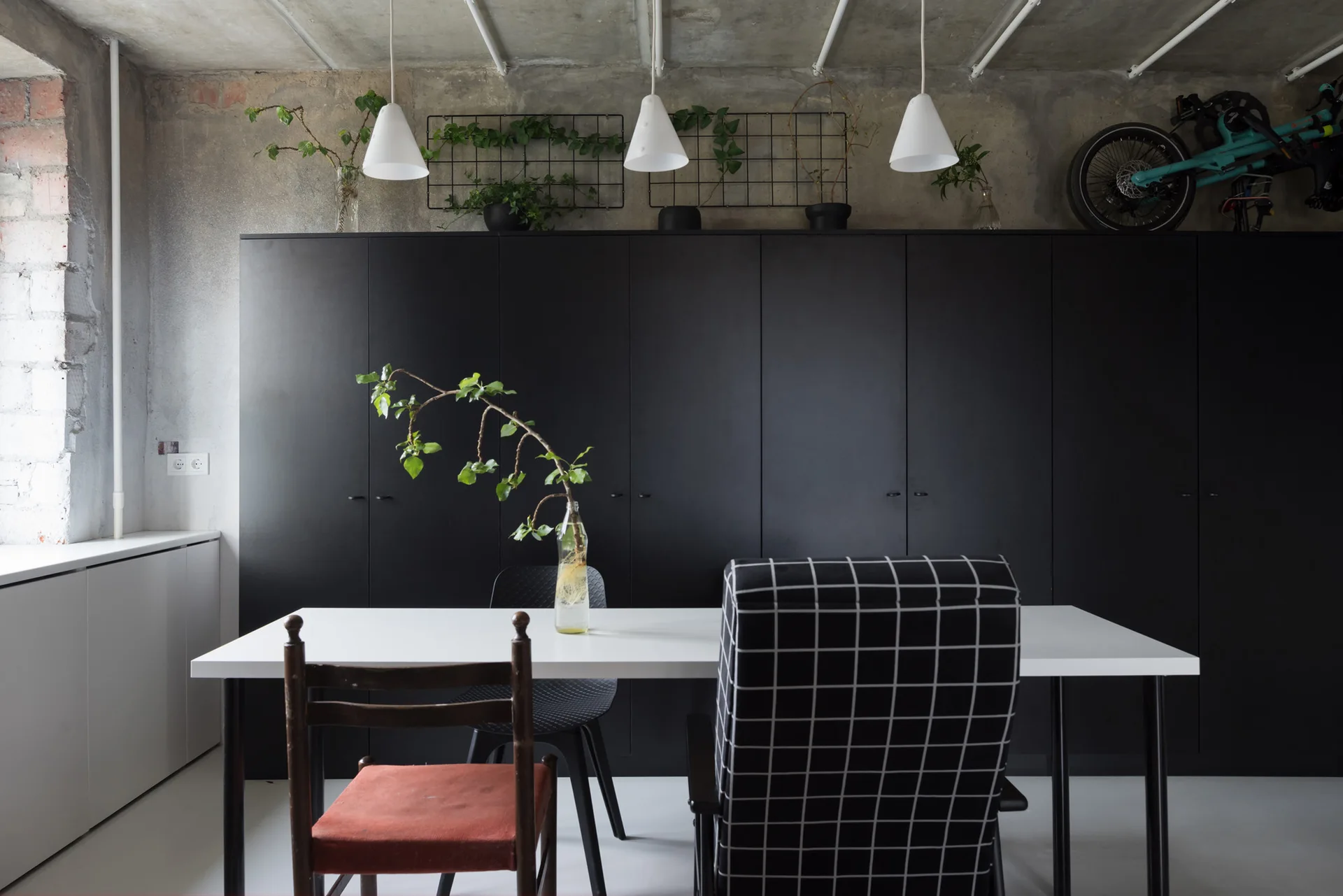 A white dining table with eclectic chairs sits in front of a floor-to-ceiling matte black cabinet against an exposed concrete wall.