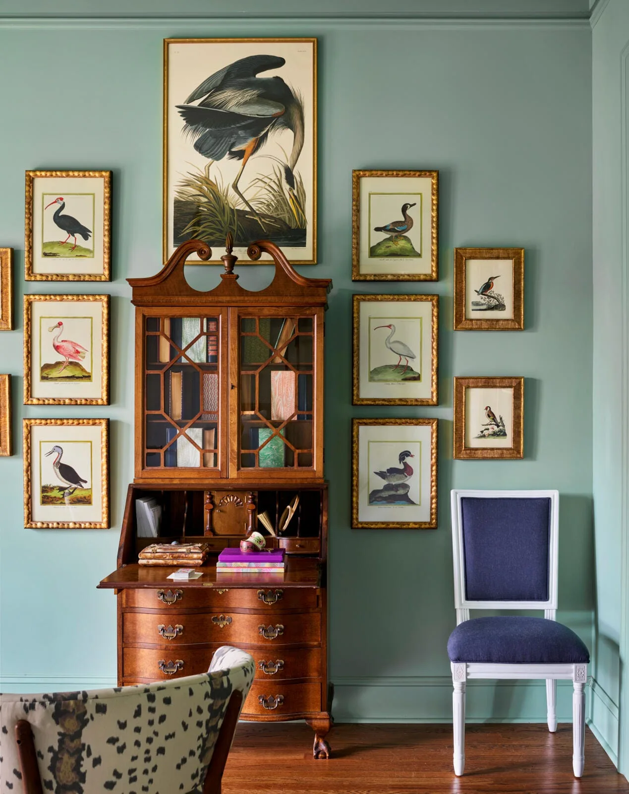 A vintage wooden secretary desk sits against a sage green wall surrounded by symmetrical rows of framed bird illustrations in gold frames.