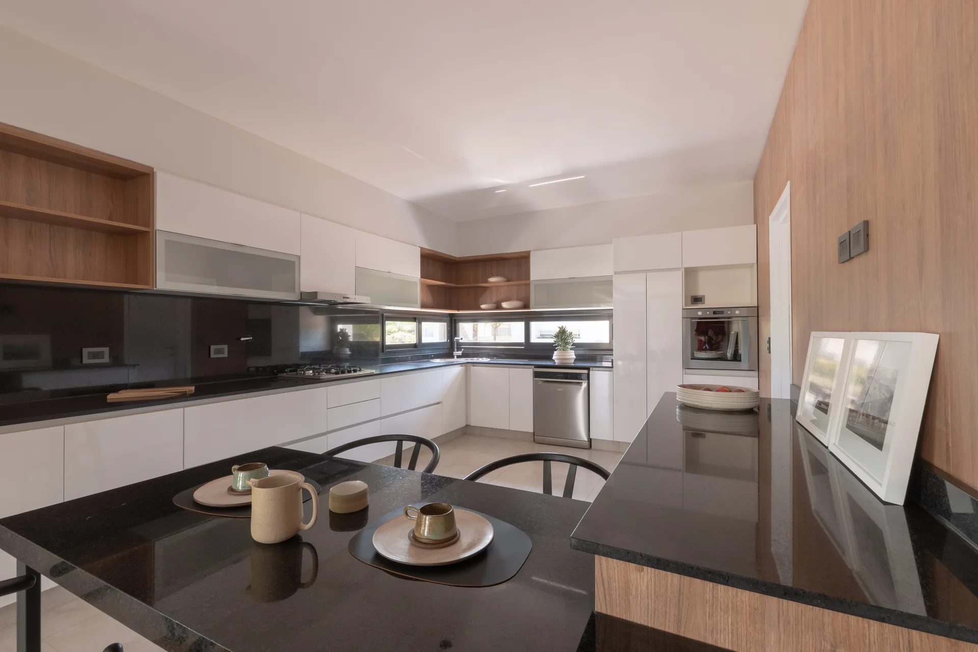 A modern kitchen featuring white flat-panel cabinets, black glossy countertops, open wood shelving, and a black dining table in the foreground.