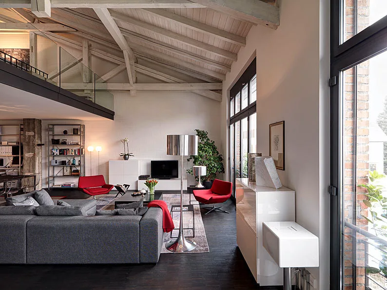 A spacious loft living room featuring a grey sectional, two red swivel armchairs, dark wood flooring, and pitched ceilings with exposed wooden beams.