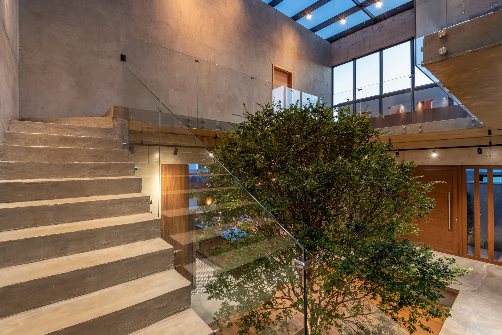 A modern concrete staircase with a glass railing ascends next to a large green tree planted inside a multi-level atrium with a skylight.