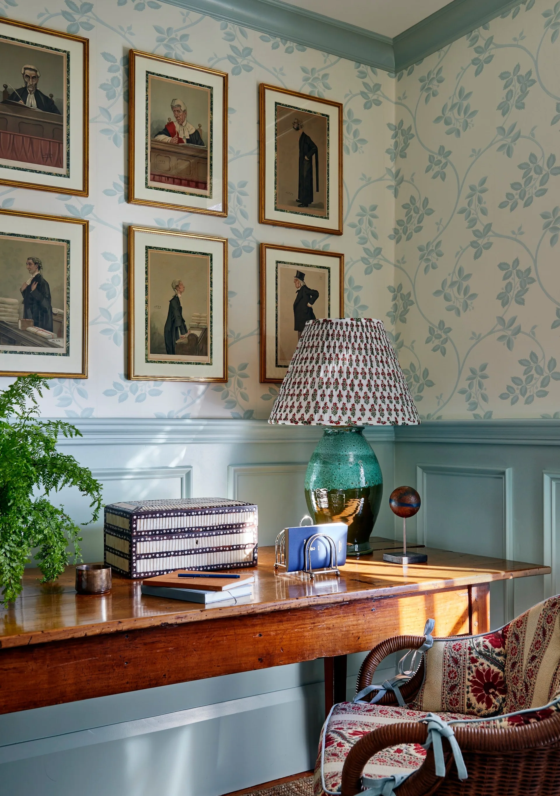 A wooden writing desk sits against light blue wainscoting and vine-patterned wallpaper, featuring a green ceramic lamp and framed portraits.