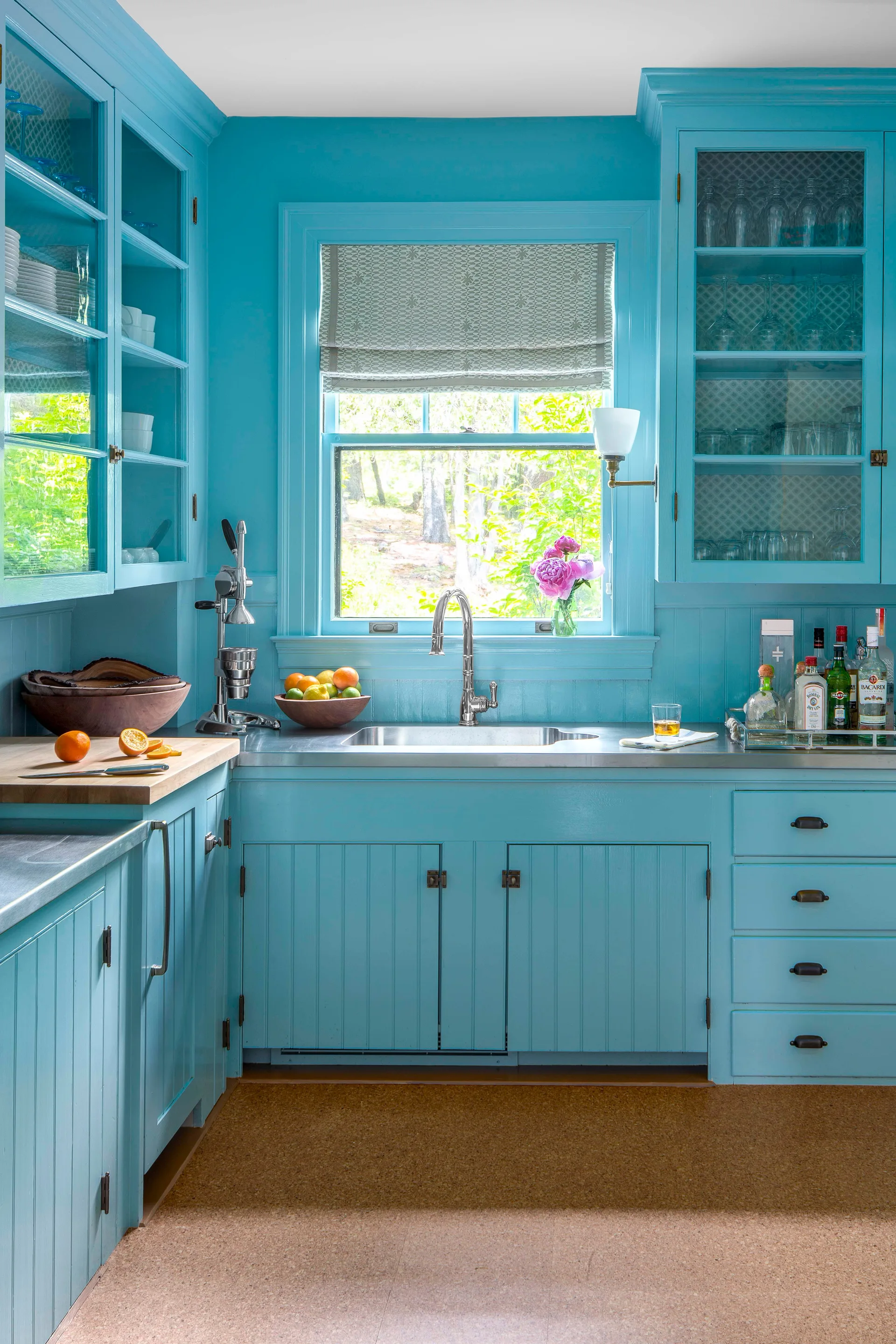 A cottage kitchen features bright blue beadboard cabinetry, stainless steel countertops, a window over the sink, and textured cork flooring.