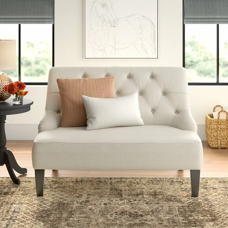 A cream tufted loveseat with orange and white throw pillows sits on a patterned rug in front of a framed horse sketch.