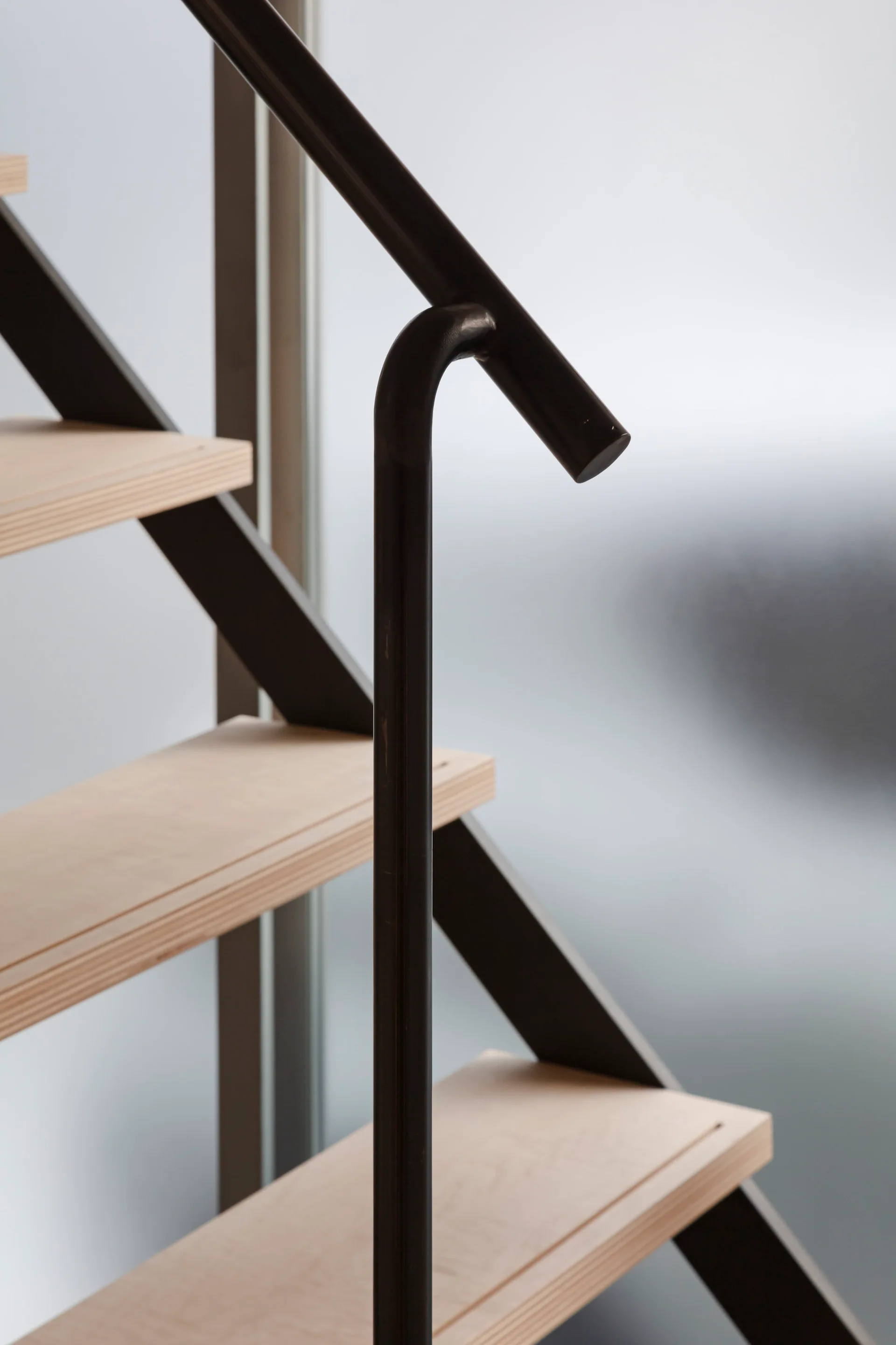 A close-up of a modern staircase featuring light wood plywood treads with exposed edges and a dark matte metal handrail.