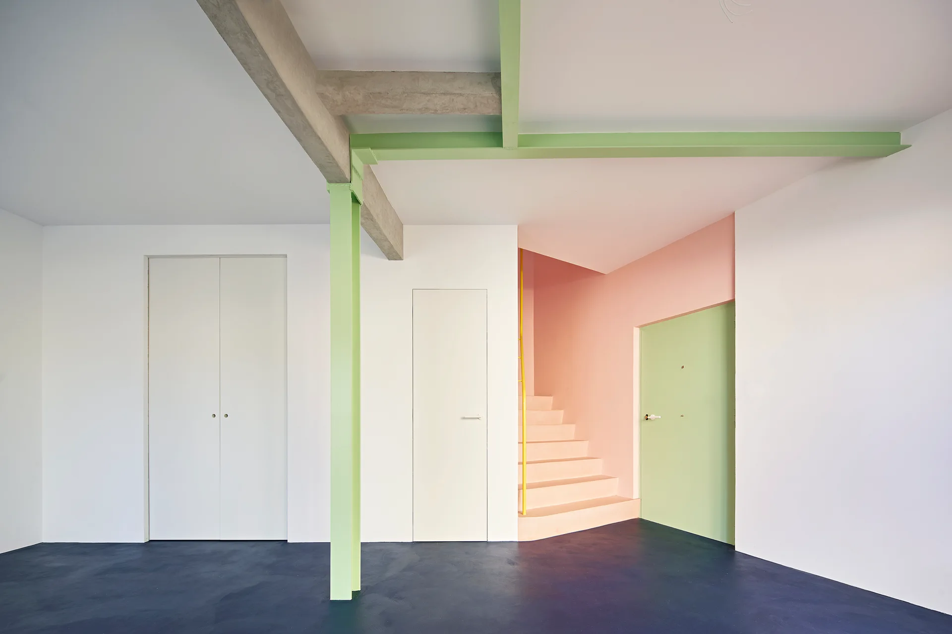 A modern minimalist interior with white walls, a dark blue floor, a light green structural column, and a soft pink stairwell.