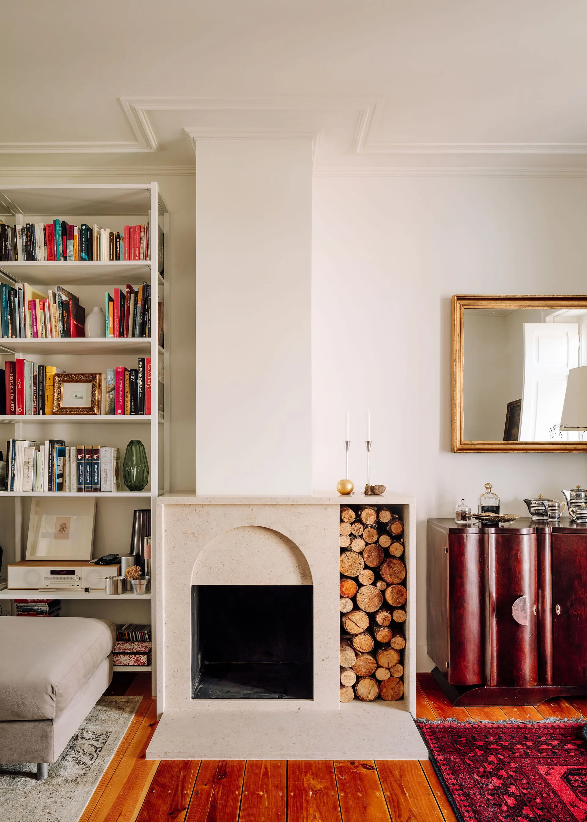 A light stone fireplace with an arched opening and integrated log storage sits between a white bookshelf and a dark wood sideboard.