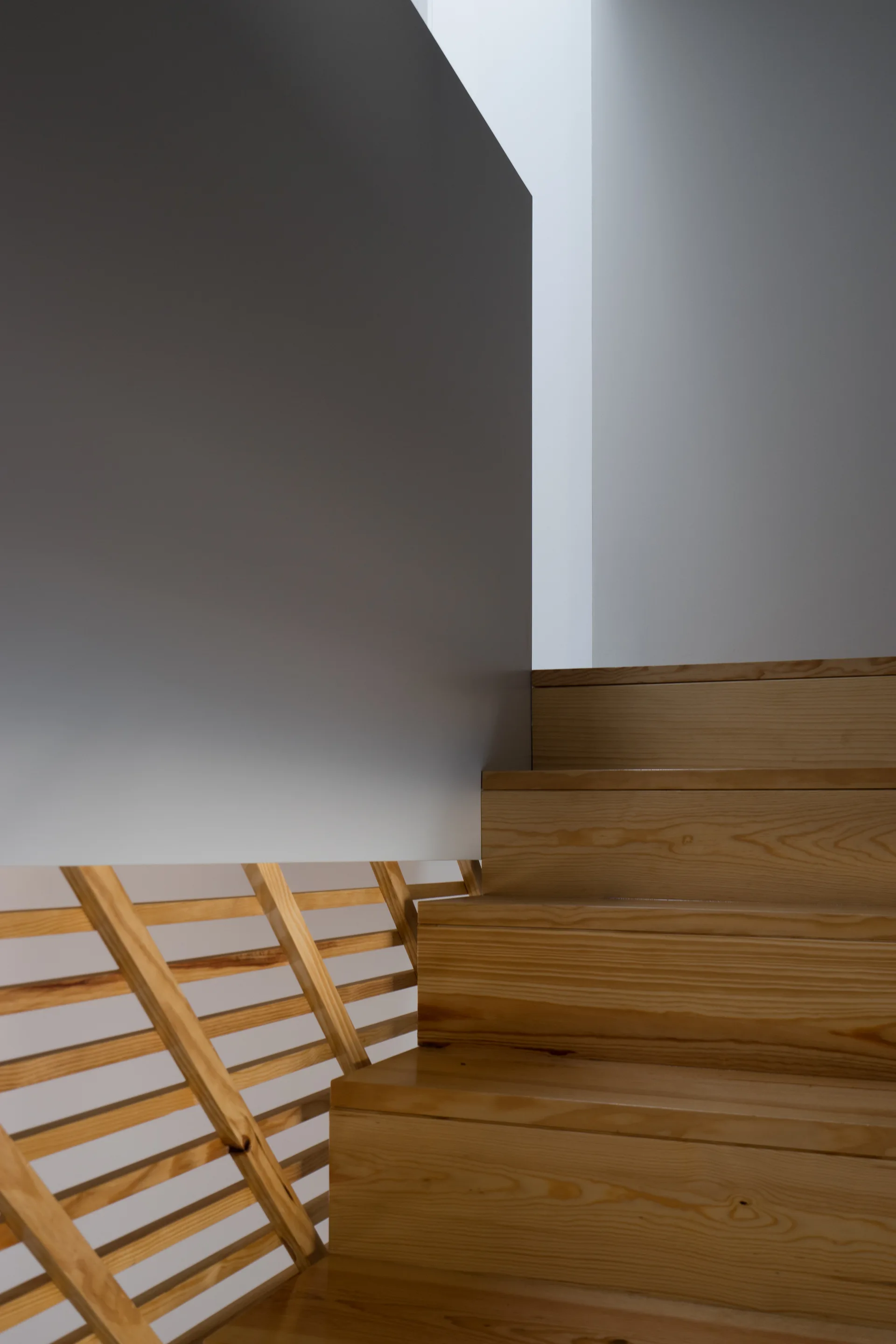 A close-up view of a modern wooden staircase with light brown steps, a solid grey wall panel, and diagonal wooden slats.