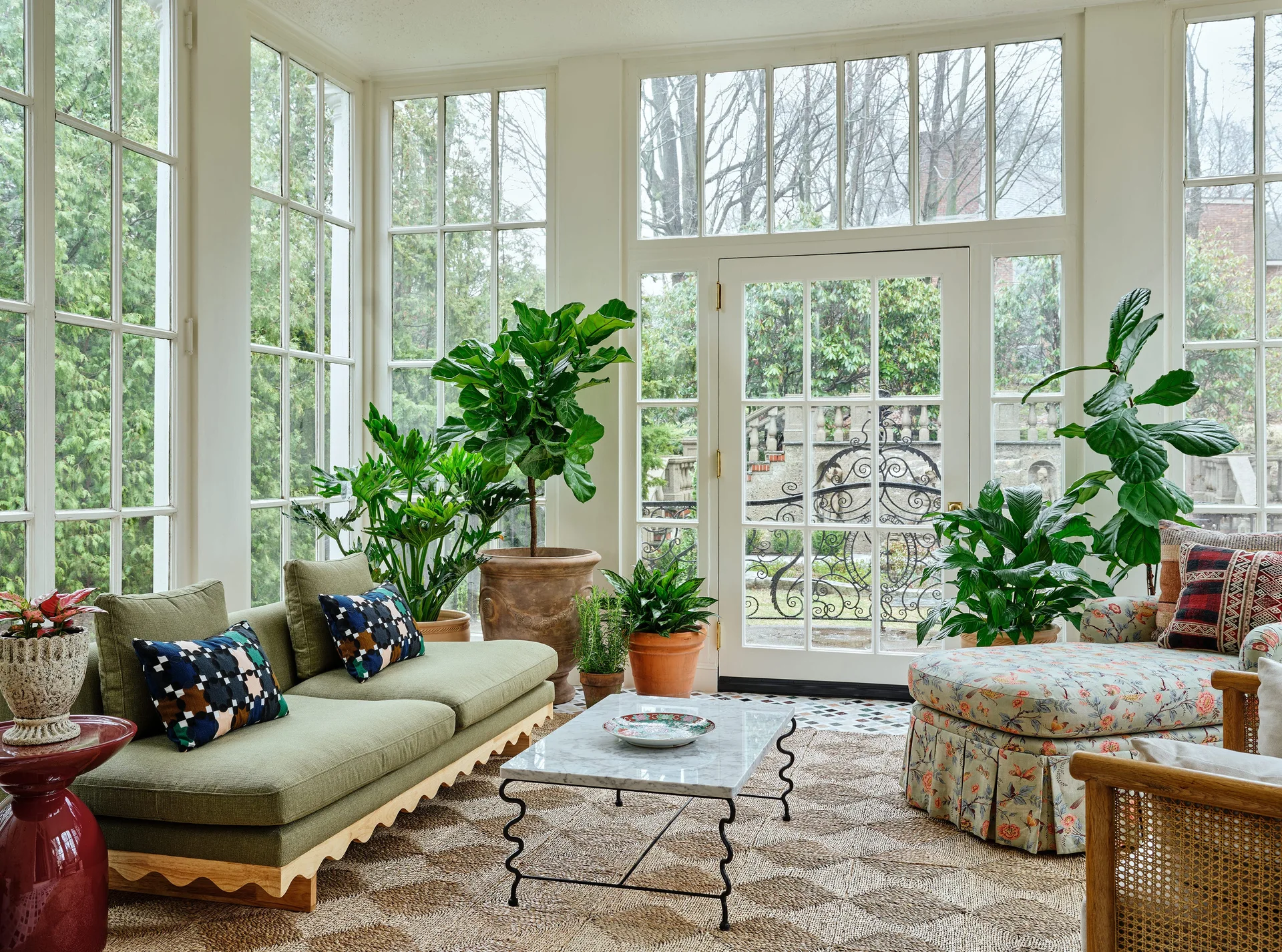 A bright sunroom features a green sofa with a scalloped wood base, a marble coffee table, and large potted plants against multi-pane windows.