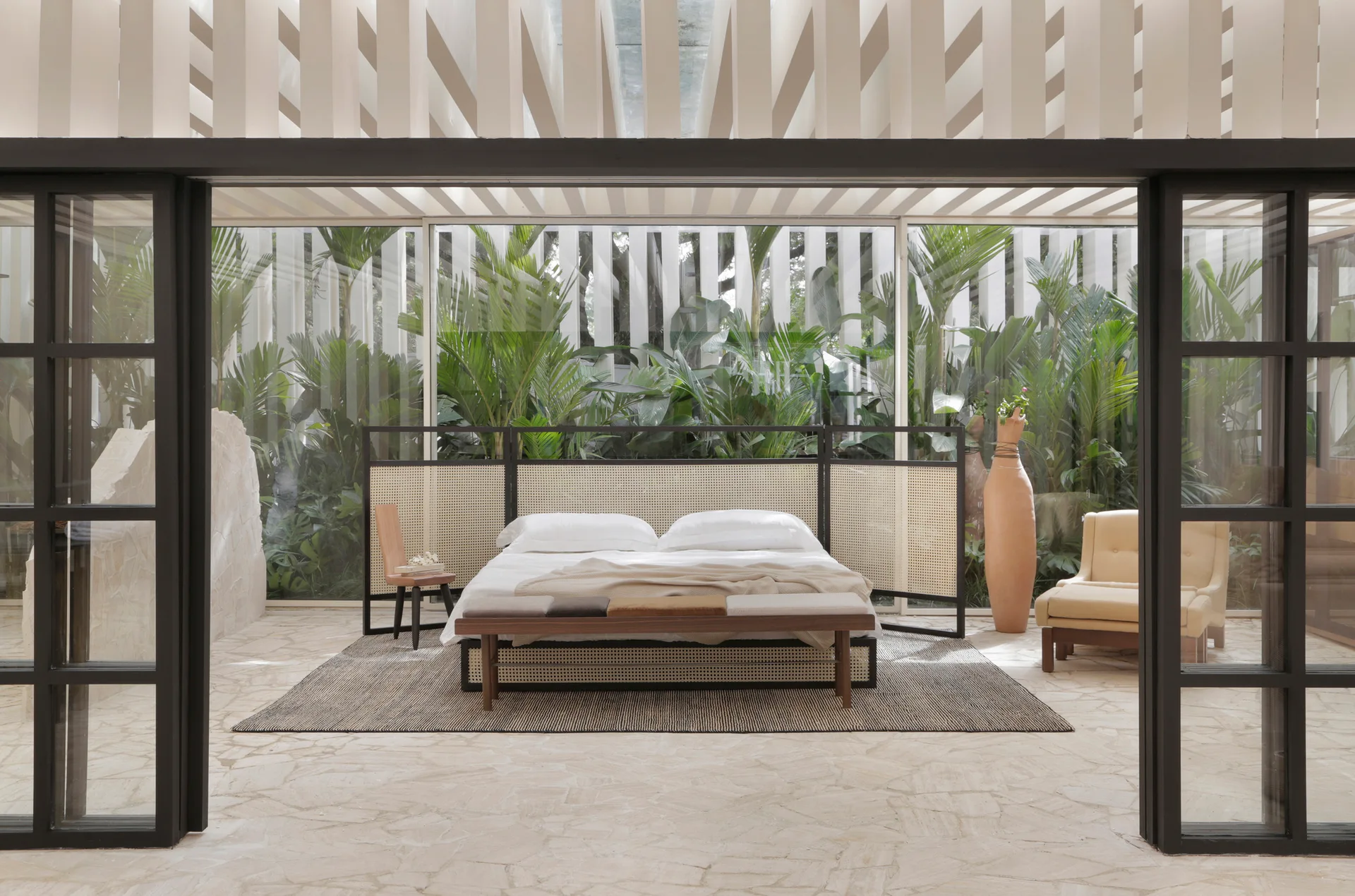 A minimalist bedroom featuring a bed with a woven cane headboard, an irregular stone tile floor, and a glass wall viewing a lush green garden.