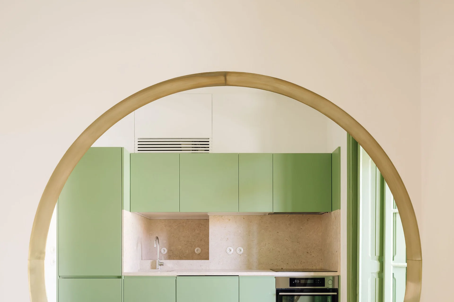 A large brass archway frames a minimalist kitchen featuring matte sage green cabinetry, a light speckled stone backsplash, and a built-in oven.